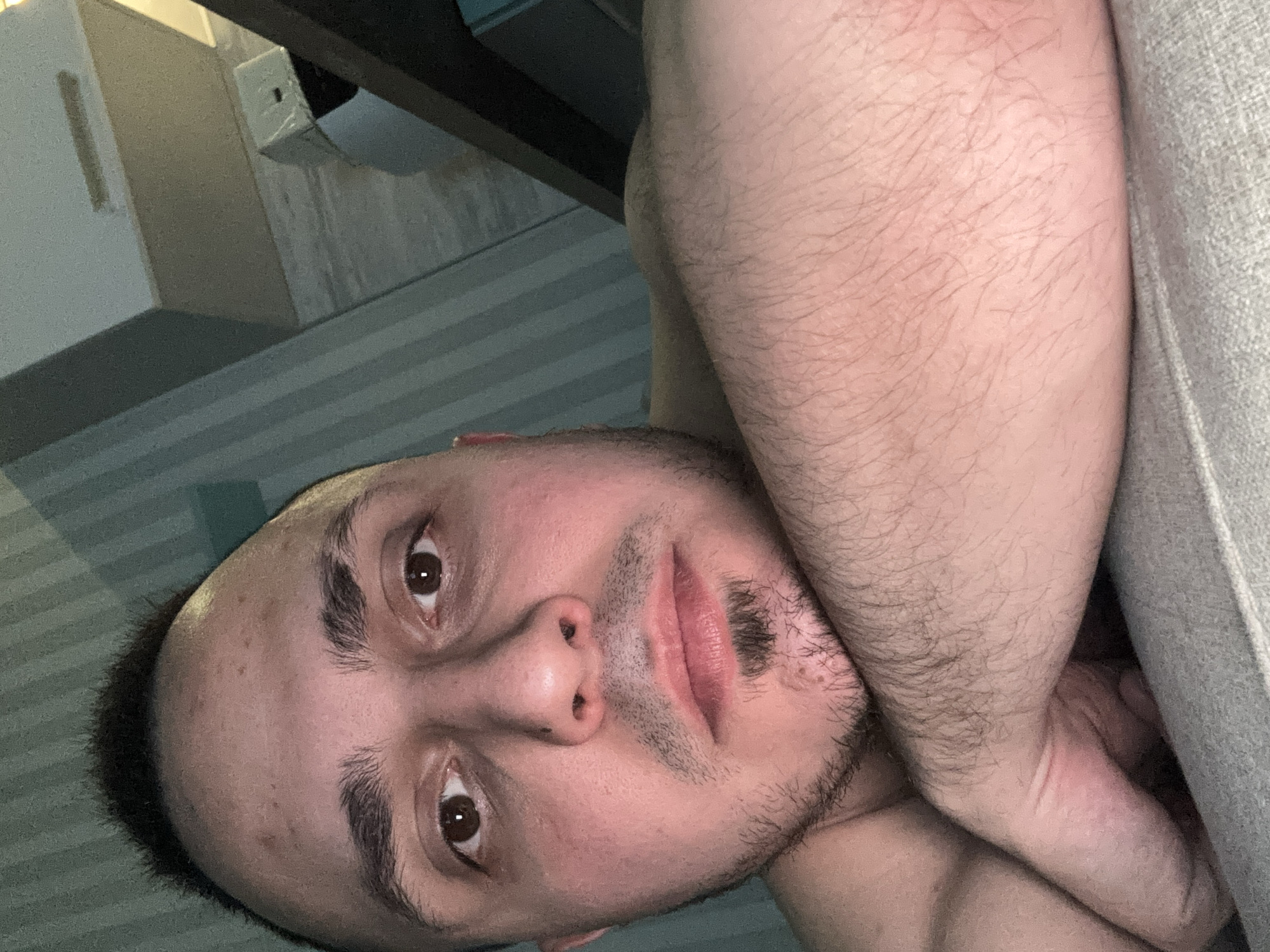 A rotated close-up portrait features an adult male from the chest up, looking directly into the camera. His head is tilted to the side, and his gaze is focused. He has dark, short hair, full dark eyebrows, and dark brown eyes. A light stubble covers his chin and mustache area. His skin appears fair with some redness, and visible body hair is present on his arms and shoulders, which are positioned across his body or resting on an unseen surface.
The setting appears to be indoors. In the background, to the left and top, are light-colored structural elements, possibly part of a wall or furniture, with subtle horizontal striped patterns. A dark, narrow bar spans the upper portion of the background. To the right of the subject, a light gray, textured fabric, likely a couch or blanket, is visible. The lighting is ambient, casting soft shadows, and does not provide specific cues about the time of day. The man's expression is neutral and engaging. There is no visible text in the image.