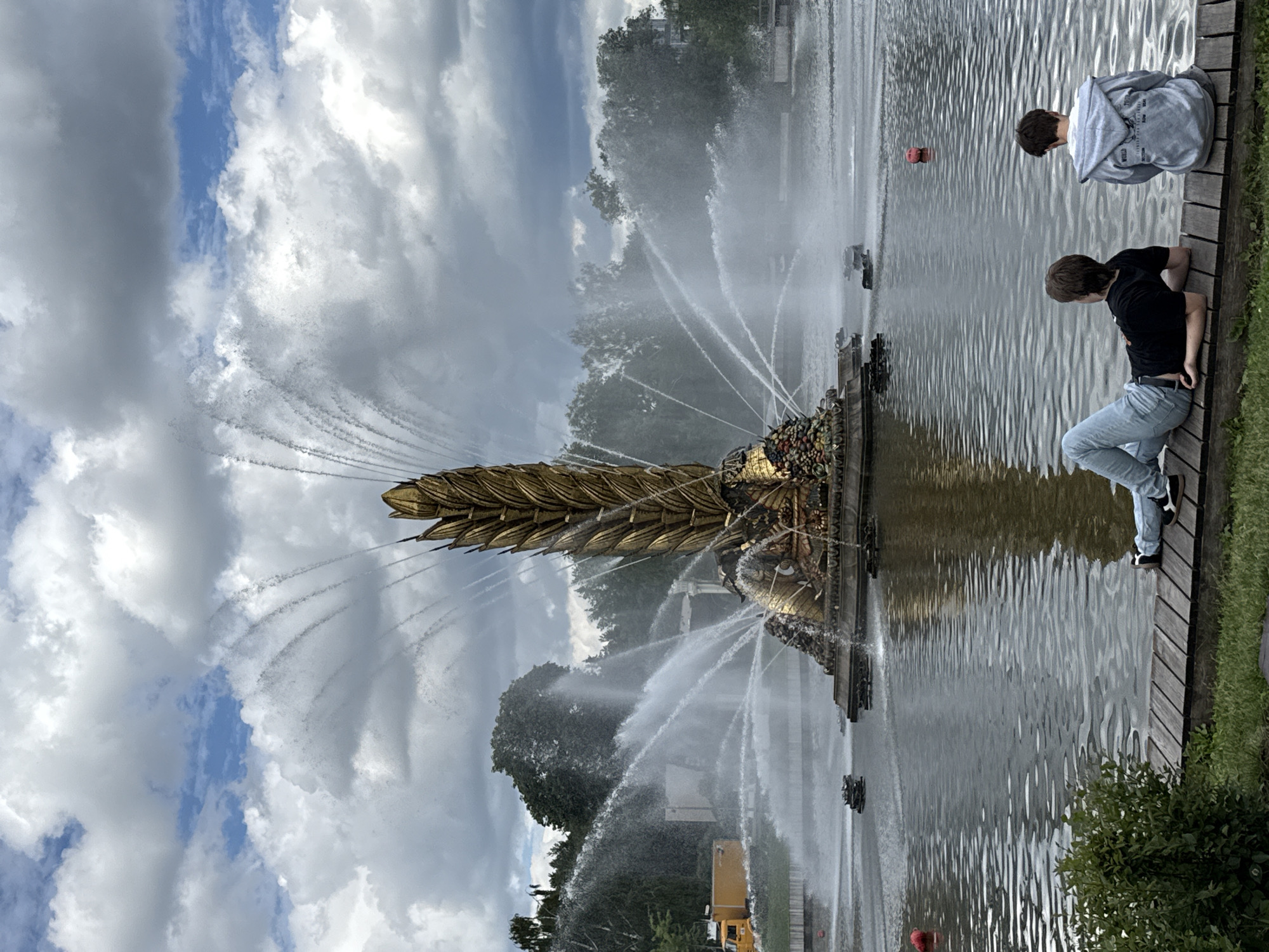 The image depicts a large, ornate fountain in a body of water. The fountain is golden in color and shaped like a massive stalk of wheat or a similar plant, with water erupting from its top and sides, creating a dramatic spray. At the base of the fountain, there appear to be sculptural elements, possibly depicting fruits or other decorative motifs.
In the foreground, two young males are seated on a wooden deck overlooking the water and the fountain. The person on the left is lying down, wearing a black t-shirt and light blue jeans. The person on the right is sitting up, wearing a light gray hooded sweatshirt and gray pants.
The environment is a park-like setting with trees visible in the background. The sky is overcast with dramatic, heavy clouds, but patches of blue sky are visible. A yellow vehicle, possibly a truck or service vehicle, is parked in the distance on the left side of the image. The water of the pond or lake reflects the sky and the fountain. The scene suggests a public outdoor space, likely a park or a leisure area in a city.
This fountain is the "Stone Flower" (Kamenniy Tsvetok) fountain, a famous landmark located in VDNKh (Exhibition of Achievements of National Economy) in Moscow, Russia.