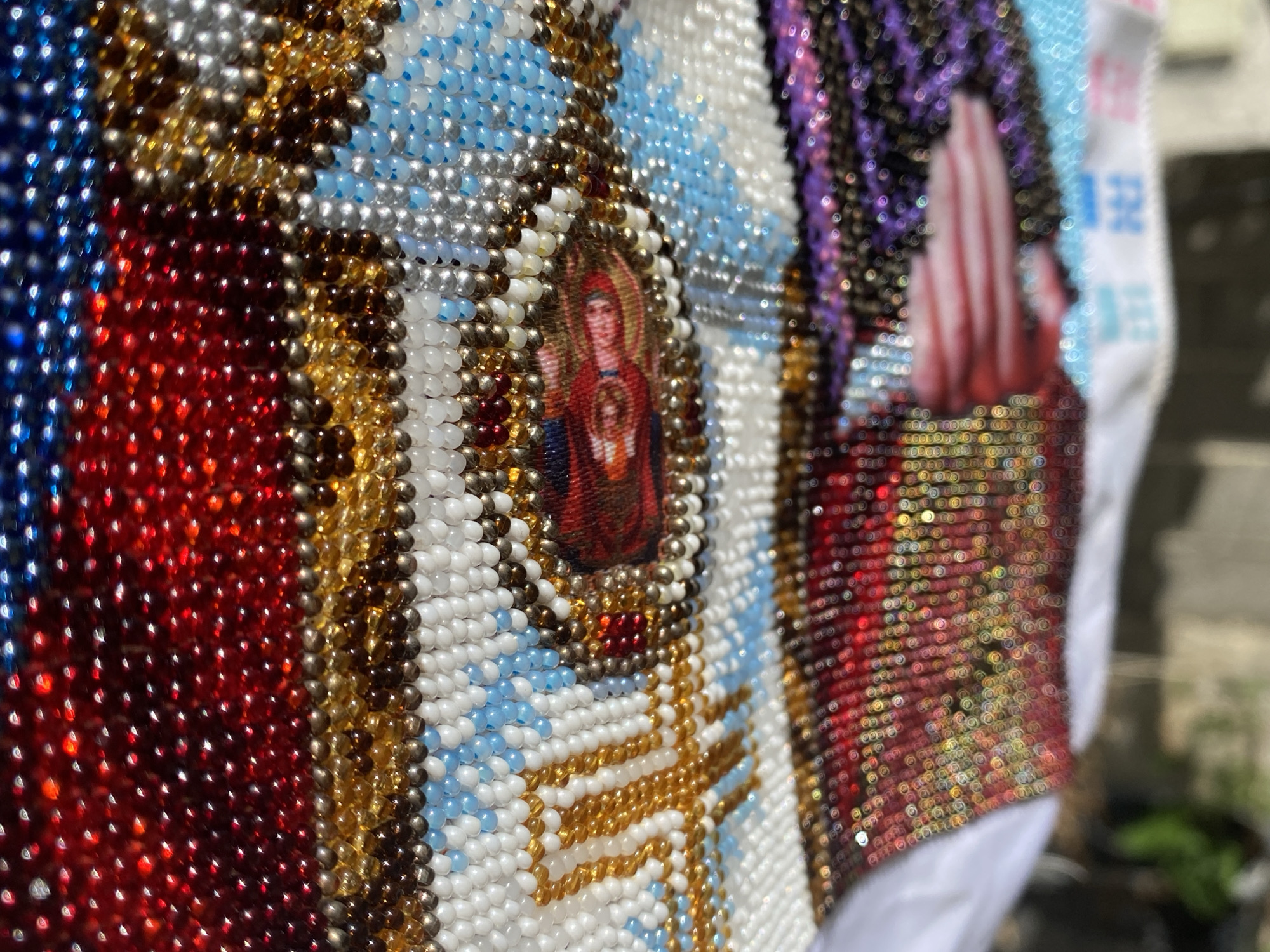 The image is a close-up shot of a heavily beaded garment, possibly a vest or a piece of clothing, with intricate patterns and a small religious image. The beads are small and appear to be glass, arranged in rows and sections of various colors, including red, blue, white, gold, silver, and purple. The religious image, centrally located and surrounded by beads, depicts a figure in red robes holding an infant. The lighting highlights the reflective qualities of the beads, creating a shimmering effect. The background is blurred, suggesting an outdoor setting with some foliage visible, hinting at an urban environment. However, there are no specific landmarks or details within the image that would allow for the precise identification of the location as Zgrade, Montenegro. The garment is the sole subject of focus, and there are no people, actions, or interactions depicted.