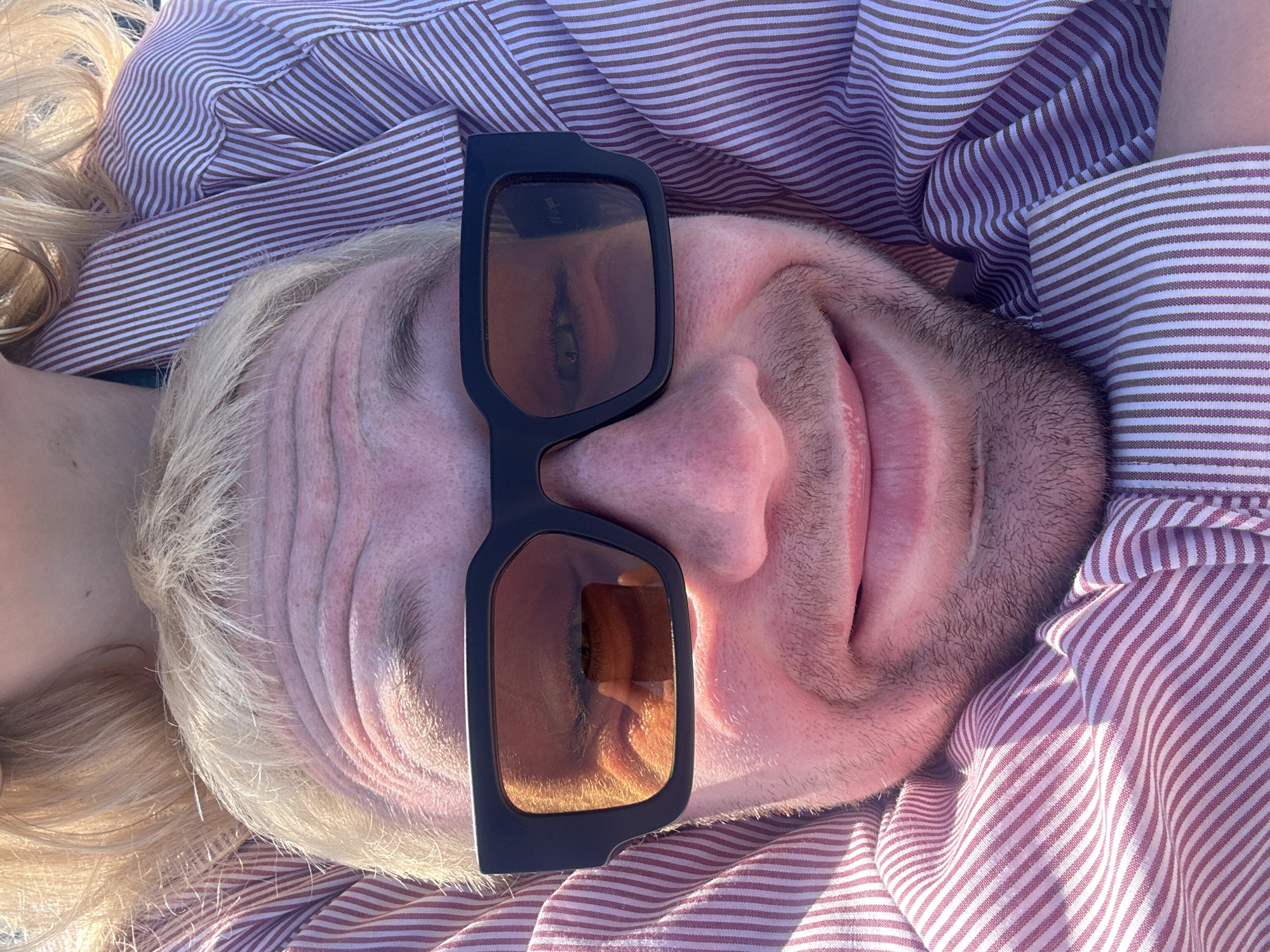 An overhead, close-up shot features a person lying down with blonde hair, fair skin, and light stubble on their cheeks and chin. The image is rotated 90 degrees clockwise, presenting the person's head on the left and chin on the right. They are wearing black-framed sunglasses with brown-tinted lenses, covering their eyes and part of their nose. A slight smile is visible around their lips beneath the sunglasses.
Reflected in the right lens of the sunglasses, a distinct eye is visible, along with what appears to be part of a hand holding a device (likely a camera or phone) and a bright sky, suggesting the perspective of the person taking the photo. Faint white text, possibly "MYRTIPS" or a similar logo, is discernible on the upper portion of the right lens. The person's blonde hair is spread out around their head.
They are wearing a collared shirt with fine, alternating light purple/mauve and white horizontal stripes, which also serves as the background. The fabric of the shirt is creased around their head and neck. The lighting is bright and sunny, casting subtle shadows and highlighting the texture of the skin and hair, indicative of daytime. The scene conveys a relaxed, casual mood, possibly on vacation in Lipari, Italy.