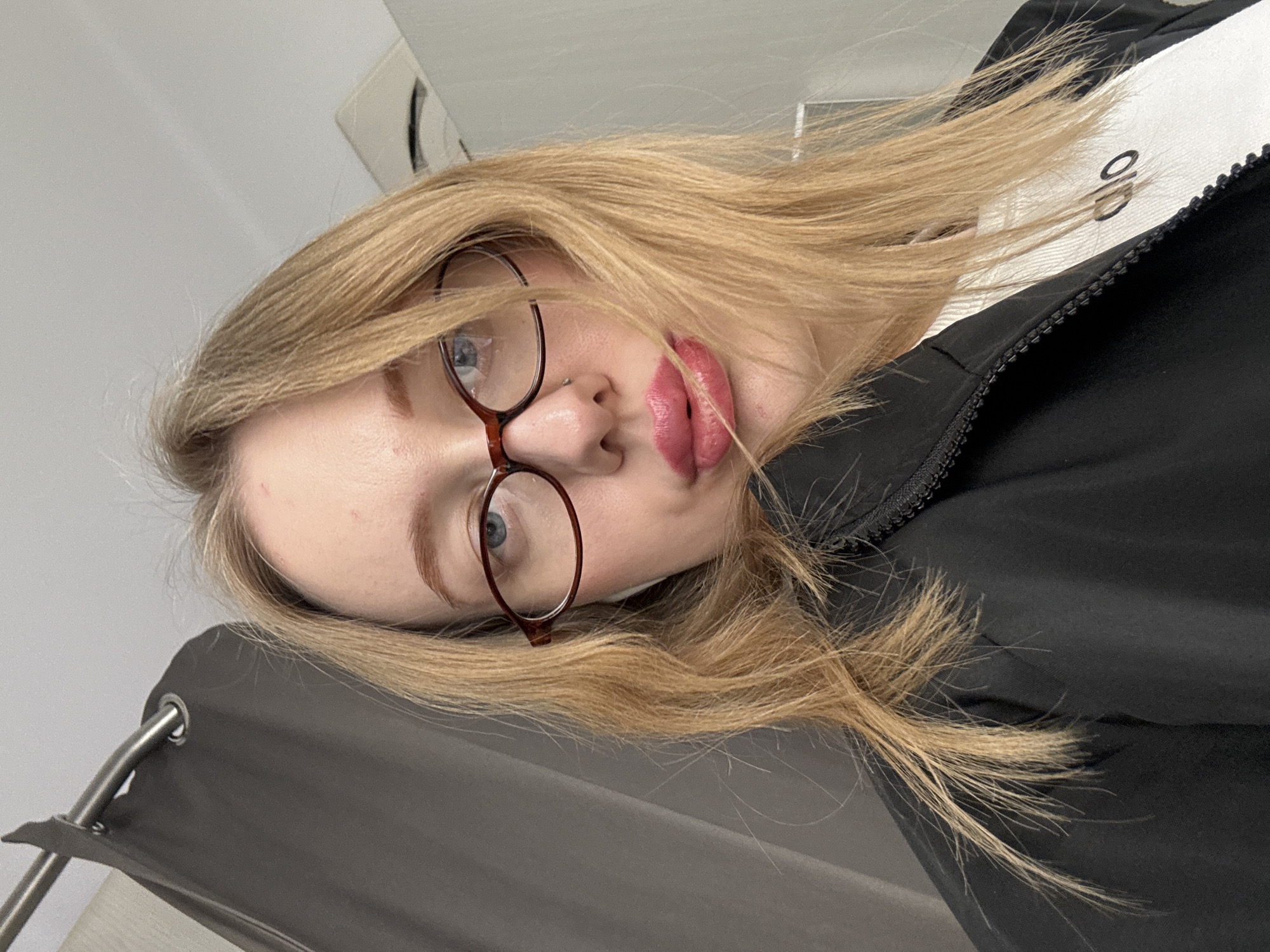 A female individual, appearing young adult, is depicted in a close-up, forward-facing selfie-style photograph. She has long, blonde hair and blue eyes, and wears brown oval-framed glasses. A small silver stud piercing is visible on her right nostril. Her lips are painted with pink lipstick and are slightly pursed. She wears a black collared jacket with a visible zipper detail over a white ribbed top, which shows a partial "alo" logo on the chest. A white wireless earbud is present in her left ear. The background on the left features a dark gray fabric curtain hanging from a silver metal rod with a grommet. The background on the right and top consists of a plain light-colored wall or ceiling, with a white rectangular ventilation unit visible in the upper right corner. The scene is an interior room. No specific visual identifiers within the image enable a determination of a precise location within Rostov-on-Don, Russia.