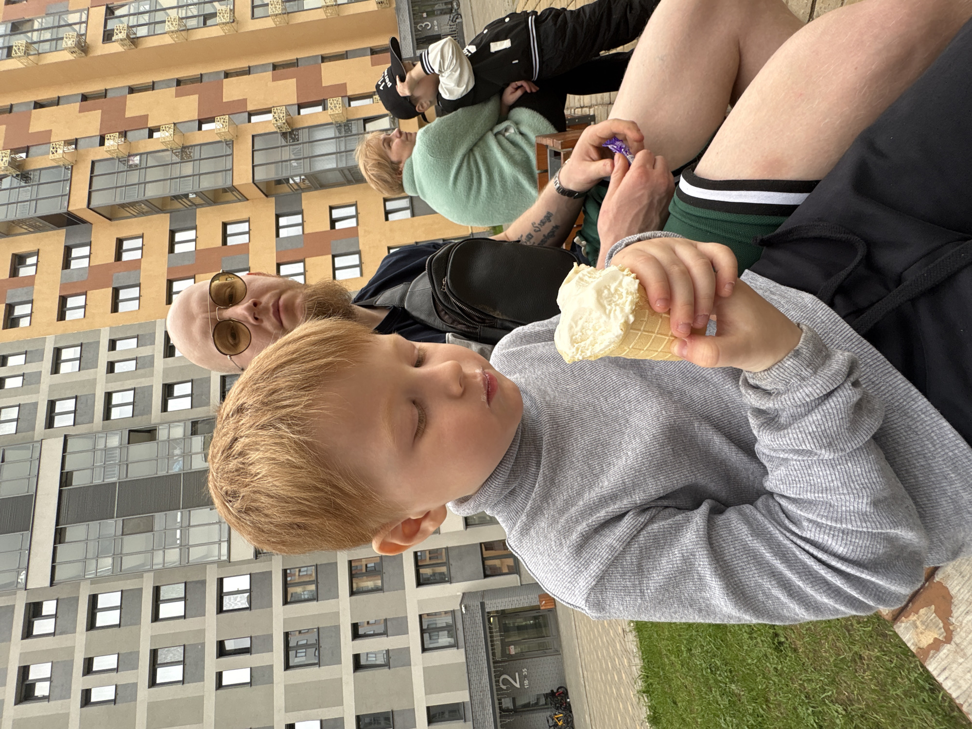 The image shows a group of people outdoors in Izhevsk, Russia. A young boy in a gray turtleneck sweater holds and eats an ice cream cone. An adult male with a bald head, sunglasses, and a beard sits beside him. Behind them, another adult sits with two younger individuals, one wearing a black and white jacket and hat. A black shoulder bag rests near the bald man. The backdrop is a multistory building with alternating gray and brown sections. A green lawn is visible at the bottom.