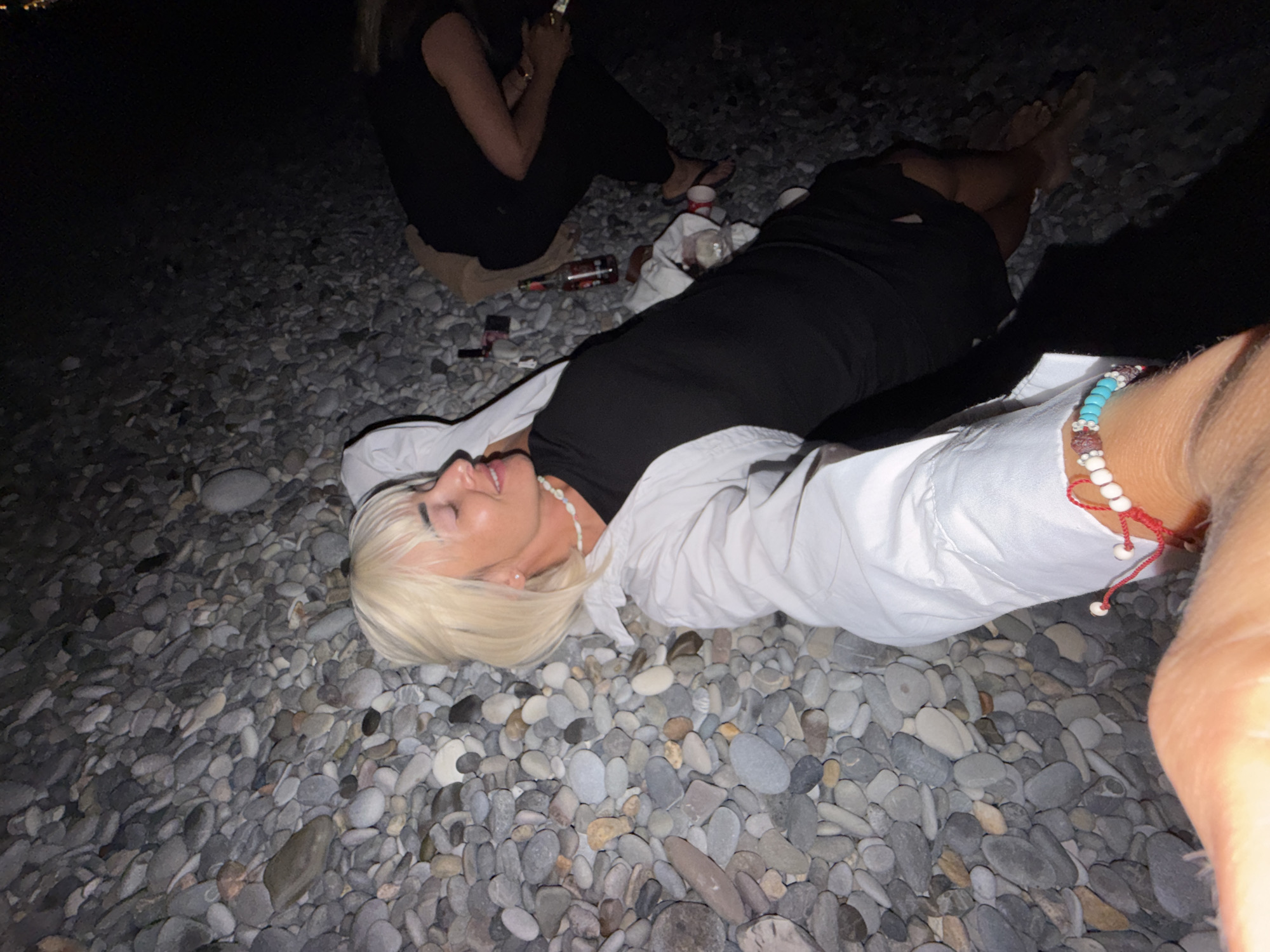 The image depicts a group of people gathered on a pebble beach at night, possibly in Sochi, Russia, as indicated by the context. In the foreground, a person with blonde hair is lying down, wearing a black dress and a white open shirt. They are looking to the side with their eyes closed and a smile on their face, adorned with a beaded necklace and a bracelet on their wrist. Another person, dressed in black, is partially visible in the background, also on the beach. Several bottles, likely containing beverages, and a few other objects are scattered on the pebbles. The lighting suggests it is nighttime, with some artificial illumination contributing to the scene. The environment is a natural beach covered in smooth, grey pebbles, with the dark expanse of the sea or a dark area of the beach in the background.
