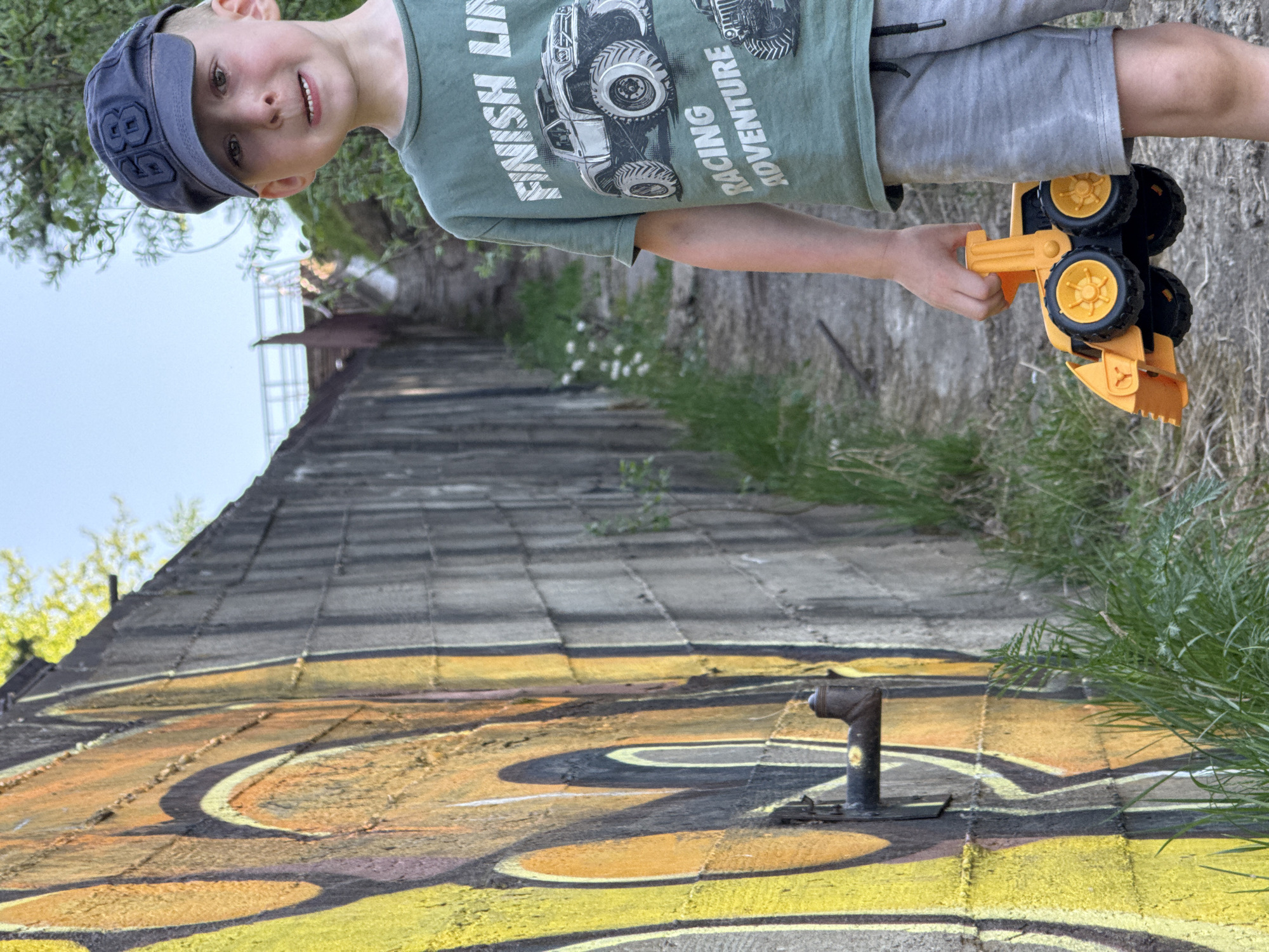 A young boy stands on a concrete structure, holding a yellow and black toy bulldozer. He is wearing a green t-shirt with a monster truck graphic, gray shorts, and a blue cap. The concrete structure appears to be a wall or ramp with graffiti and a metal pipe sticking out. There is vegetation growing alongside the structure. The location is most likely in a park or similar public area. The image is taken from a low angle. I am unable to identify the exact location within the city of Noviy, Russia.
