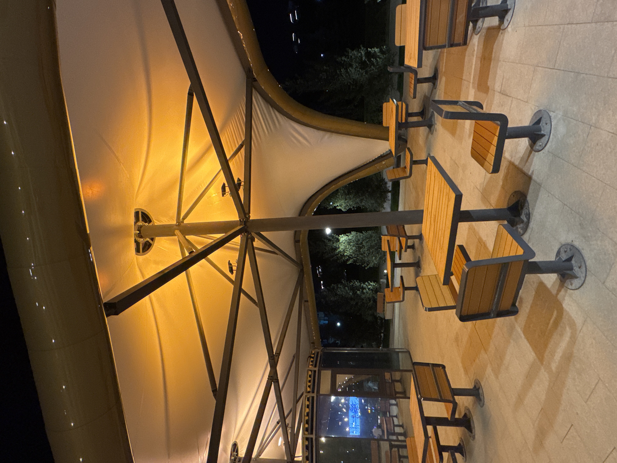 An outdoor seating area for a fast-food establishment is depicted at night, illuminated by artificial lighting. The seating area is covered by a large, white tensile canopy structure with a prominent central support pole and a radial bracing system. Tables and benches, constructed with wooden seats and backrests and dark metal bases, are arranged throughout the space. In the background, trees are visible against a dark night sky, with distant city lights suggesting an urban setting. The paving under the seating area consists of light-colored square tiles. The overall impression is of a clean, modern, and functional outdoor dining space.