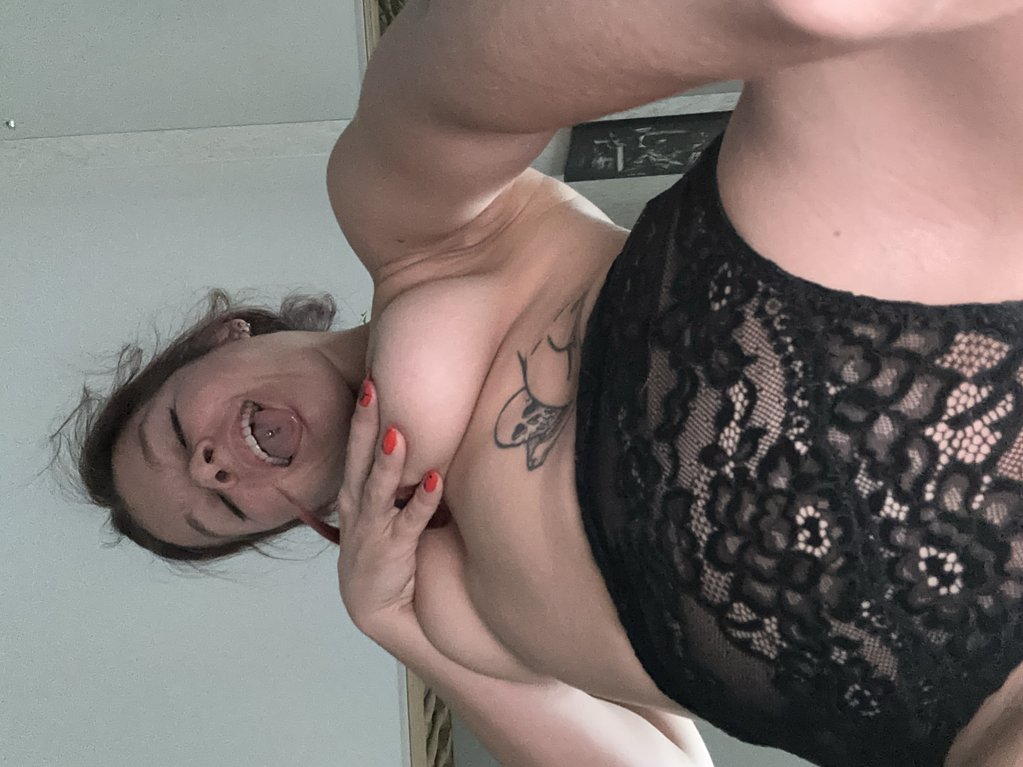 The image shows a person with dark hair and fair skin in a low-angle, upside-down shot. The person is wearing black lace underwear. They have a tattoo on their chest and a tongue piercing. Their eyes are closed, and their mouth is open, with their tongue sticking out, revealing the piercing. Their right hand is touching their breast. The background is a plain, light-colored wall. The overall lighting suggests an indoor setting, possibly a bedroom, with soft light. The emotion conveyed is one of playfulness or seduction. There is no visible text in the image. The location context provided is Vladimir, Russia.