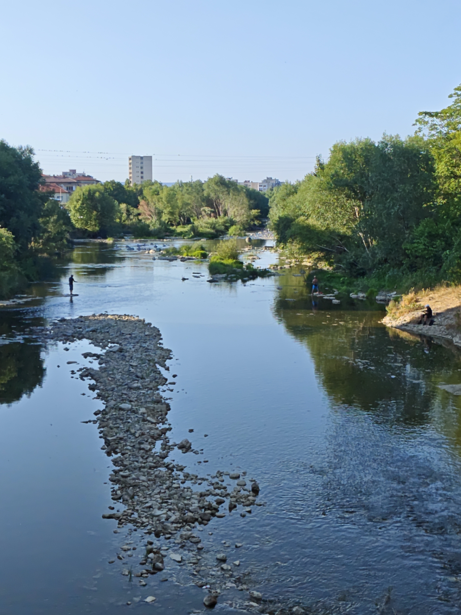 The image shows a wide, calm river flowing through a city. On the left bank, a person is standing in the shallow water, facing away from the camera. A rocky shoal extends into the river from the left bank. On the right bank, several people are visible, one sitting and two standing near the water. Trees line both banks, and in the background, buildings and a tall, slender tower can be seen, indicative of an urban environment. The sky is clear and blue. This scene depicts the Osam River flowing through the city of Lovech, Bulgaria, with the distinctive Lovech High Tower visible in the background.