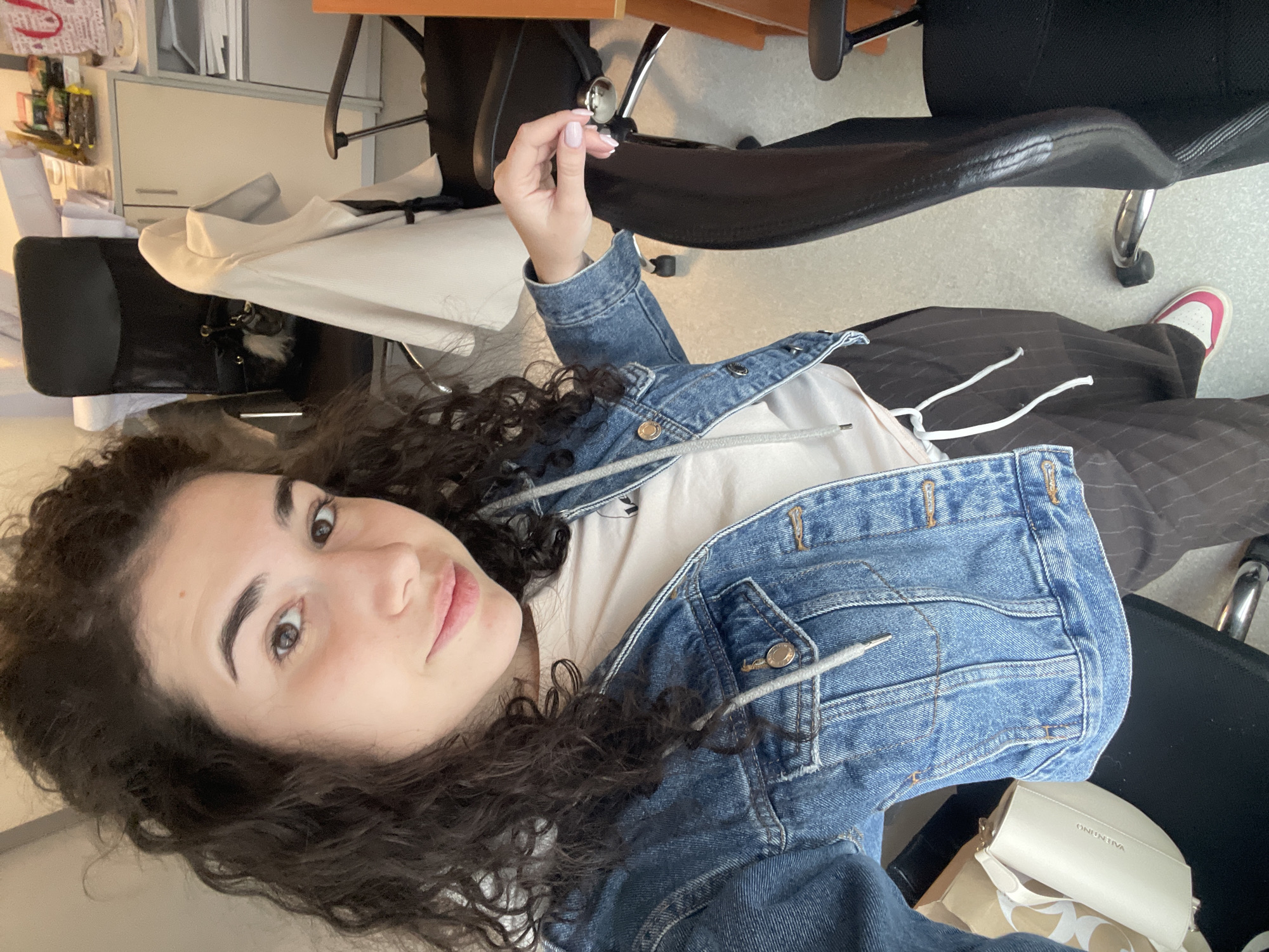 A young woman with curly dark hair is taking a selfie indoors. She is wearing a denim jacket over a light-colored top and striped pants. Her left foot, wearing a white shoe with a pink accent, is visible. The environment appears to be an office setting, with office chairs and furniture visible in the background. The location is Sofia, Bulgaria.