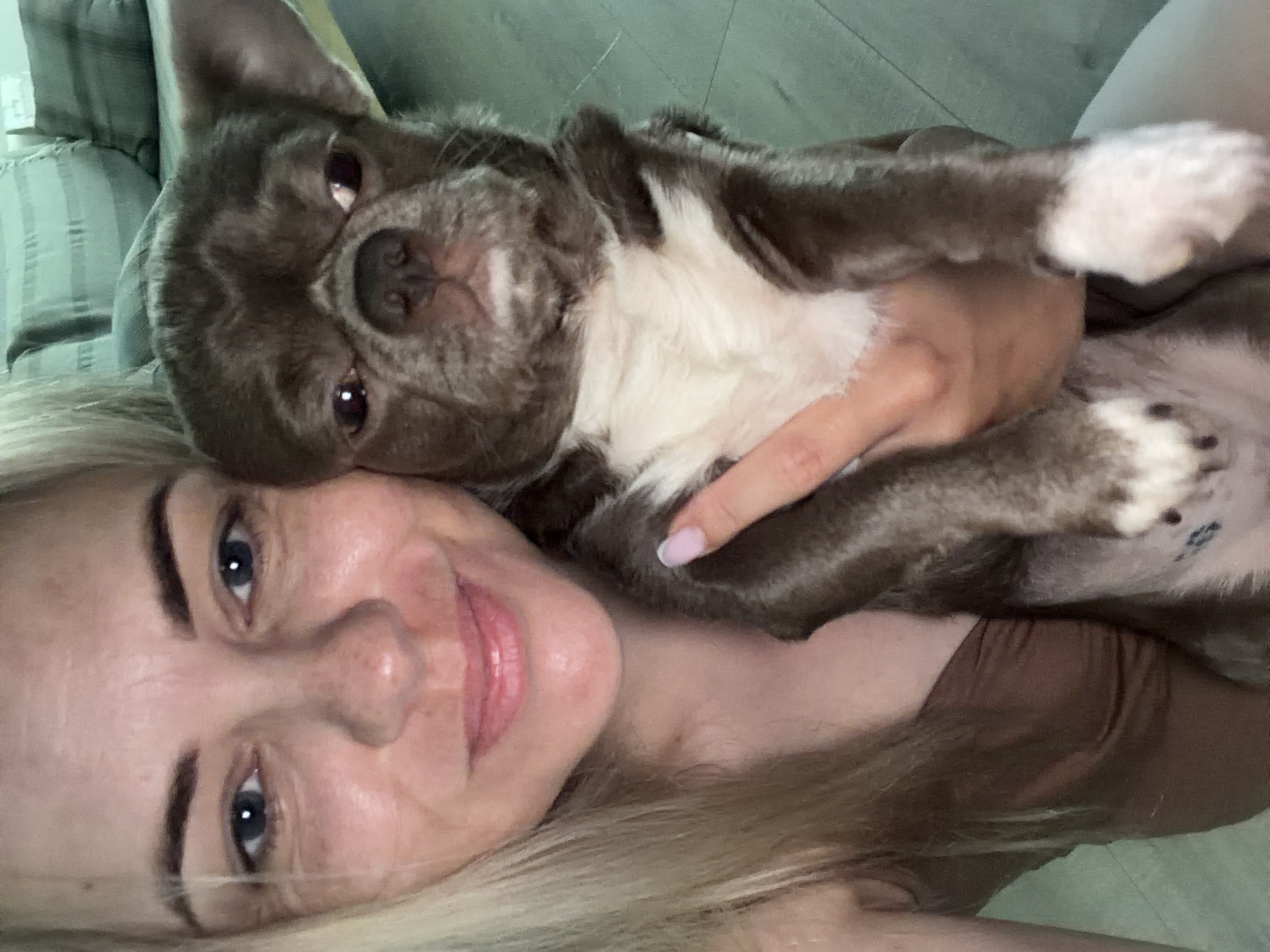 A woman with blonde hair and blue eyes is lying down, holding a gray and white French bulldog. The dog is resting on her chest, looking directly at the camera. The woman is smiling. The location is not identifiable as Votkinsk, Russia, based on the provided image.