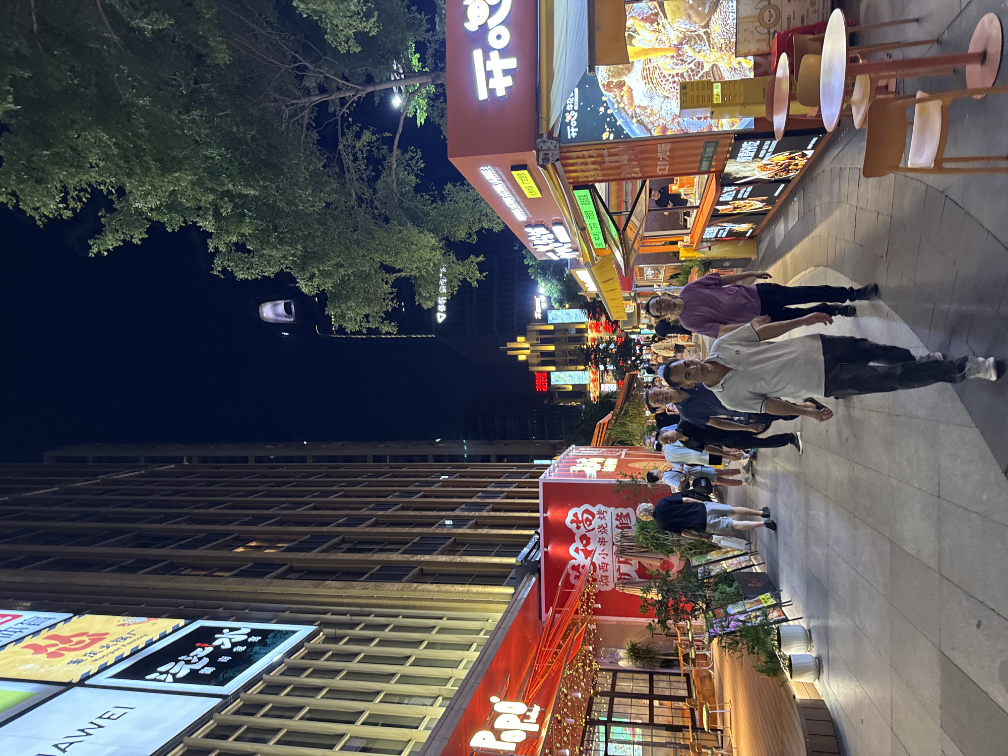 The image depicts a lively night scene on a pedestrian street in Haizhu District, China. The street is lined with brightly lit shops and food stalls, creating a vibrant atmosphere. On the left, a tall building with numerous windows is illuminated, with large signs displaying Chinese characters and the "Huawei" logo. In the foreground, a red awning extends over an establishment labeled "Popo," with tables and chairs set up outside. Further down the street, food stalls are set up in shipping containers, offering various cuisines. People are seen walking along the street, some looking at the stalls, and others engaged in conversation. The ground is paved with stone tiles, and the sky is dark, indicating it is nighttime. The overall impression is of a bustling commercial and entertainment district.