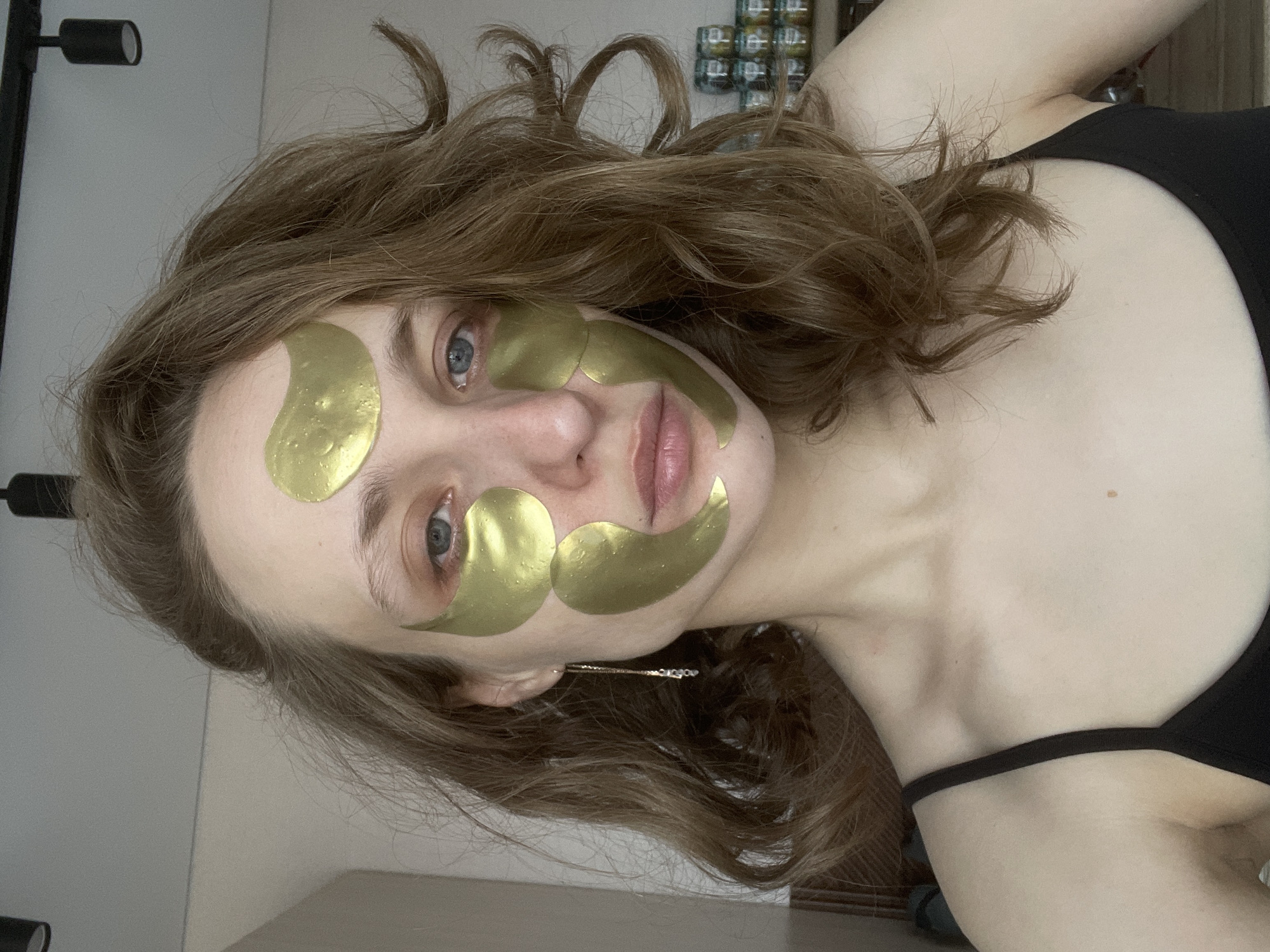A light-skinned woman with light brown, wavy hair is shown from a close, angled perspective. She is wearing several golden-colored cosmetic masks on her face and a black spaghetti-strap top. The background includes a wall with a shelf or cabinet on it and a dark fixture. There is no information in the image to confirm its location in the city of Подшивалово, Russia.
