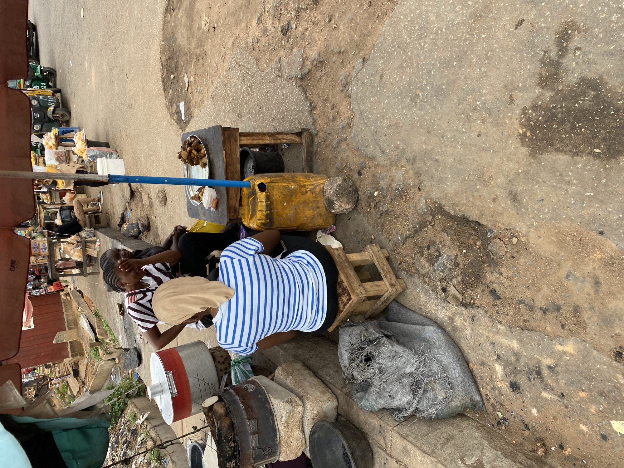 In Bukuru, Nigeria, a street food vendor operates on a paved, worn roadside. Two women are present; one wears a striped top and sits on a small, wooden stool, while the other wears a striped shirt and holds her hands near her face. The vendor’s setup includes a wooden table laden with fried food on a metal plate, resting next to a yellow plastic container. Cooking equipment such as pots and a mesh bag are also visible. In the background, motorized tricycle vehicles are parked, and a bustling market environment is suggested.