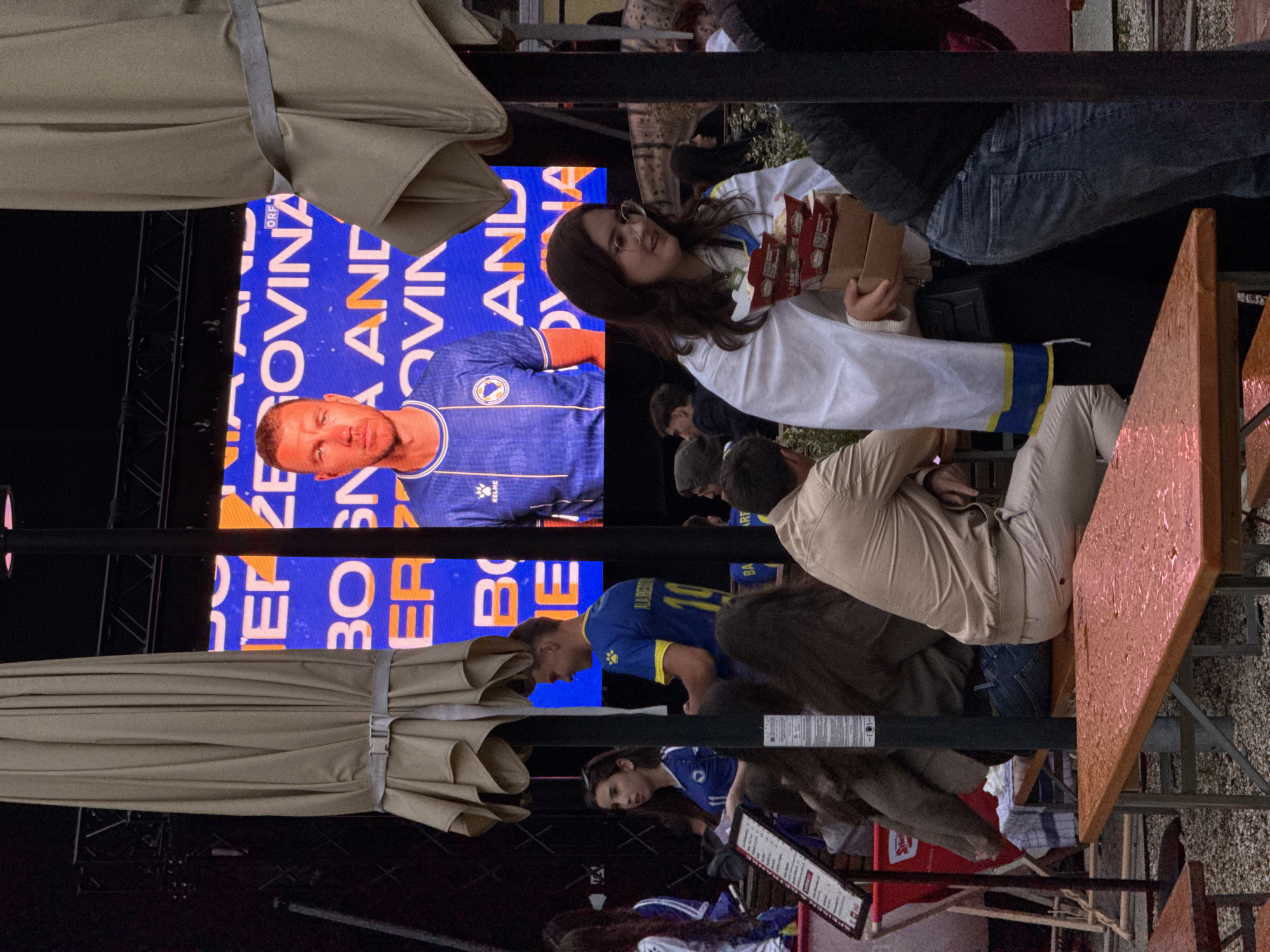The image depicts an outdoor gathering, likely a public viewing event, in front of a large screen displaying a football match. The screen shows a close-up of a male football player in a blue jersey, with text indicating "BOSNIA AND HERZEGOVINA" and "ORF". People are seated at tables in the foreground, some of whom are wearing team colors or draped in flags. One woman in the foreground, wearing glasses and a white cape with blue and yellow trim, is holding several food containers. The atmosphere appears casual and festive, with umbrellas providing shade. The presence of the ORF logo suggests the event is likely taking place in Vienna, Austria, as ORF is the Austrian public broadcaster.