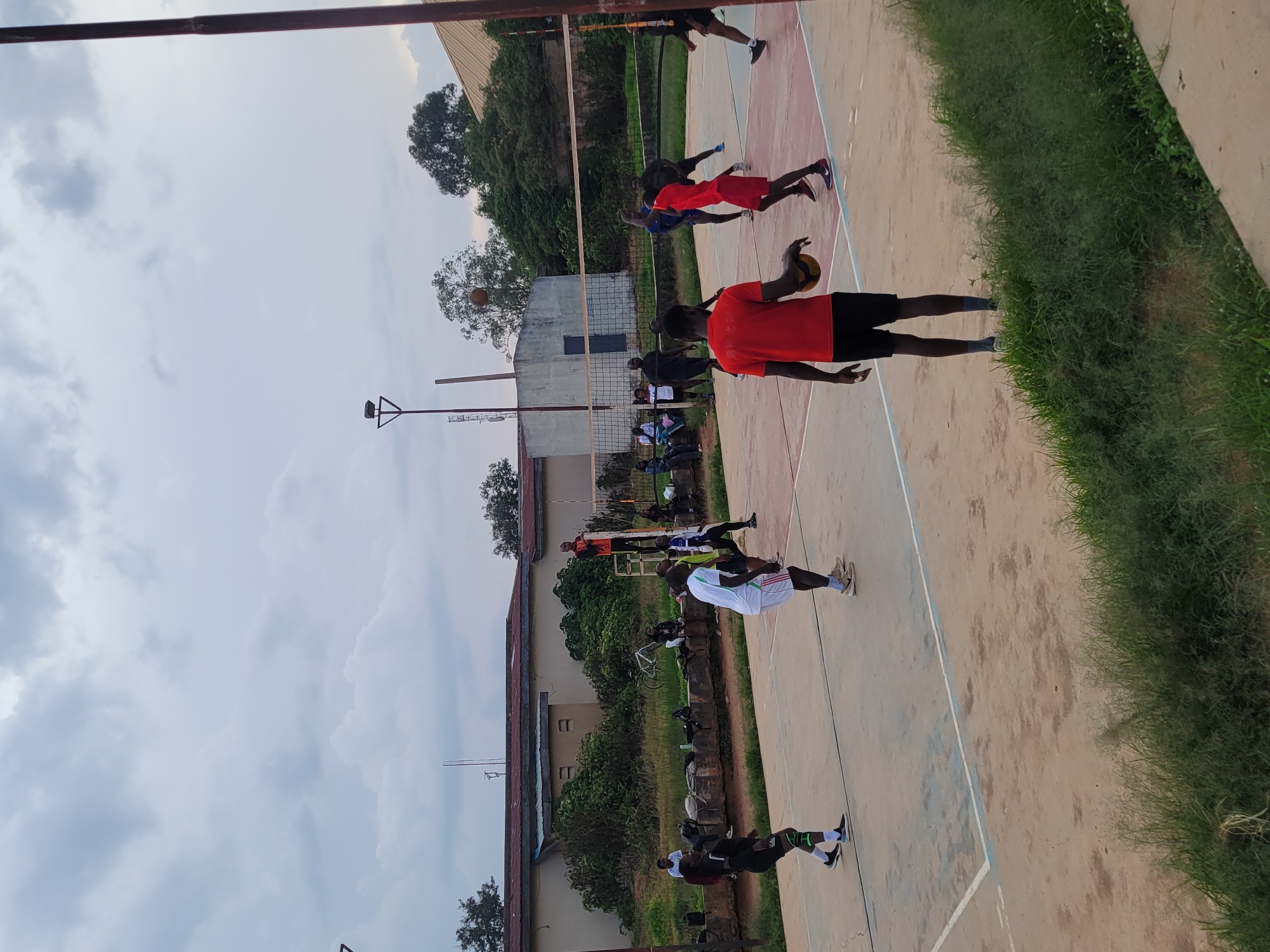 A group of people are playing volleyball on an outdoor court. The court is made of concrete with painted lines. There is a volleyball net stretched across the middle of the court. In the background, there is a building and trees. The sky is overcast. Several individuals, predominantly male, are actively engaged in the game, positioned on both sides of the net. Some players are in motion, either jumping to hit the ball or in defensive stances. One player in a red shirt and black shorts is holding a volleyball, facing away from the camera. Another player in a white shirt and white shorts is mid-action on the other side of the net. Spectators are visible sitting on a low wall in the background, and some are standing near the court. The environment suggests a sports facility or a recreational area.