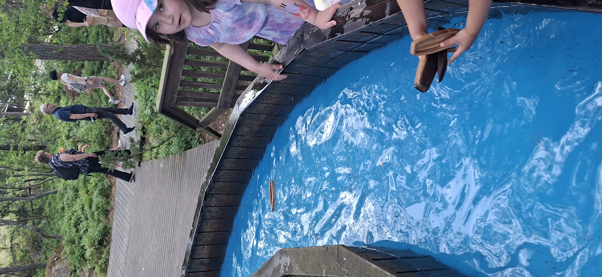 The image depicts a scene at Moominworld, an amusement park located on the island of Kailo in Naantali, Finland. In the foreground, a young girl with a pink hat and a tie-dye dress is reaching towards a wooden trough filled with bright blue water. She has a temporary tattoo of two figures on her left arm. Next to her, another child is holding two wooden boat-like toys. The trough is lined with dark wooden planks. A single wooden toy boat is visible floating on the water's surface. In the background, several people are walking on a wooden boardwalk amidst lush green foliage and trees. One woman is wearing a black backpack, and a man is wearing a blue shirt. The overall setting suggests an outdoor recreational area with a focus on water-based activities.