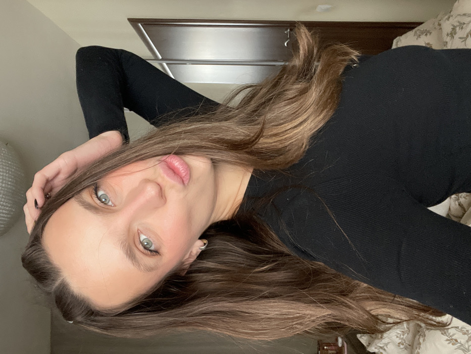 The image displays a close-up portrait of a young adult female captured indoors. The female possesses long, wavy brown hair with lighter strands, light-colored eyes, and fair skin with visible blush on her cheeks. She is dressed in a black, long-sleeved, ribbed garment. Her lips are pursed in a kissing expression, and her left hand is positioned with fingers lightly touching the top of her head. The background consists of light-colored, possibly beige, walls. To the left, a section of a bed covered with a light-colored, patterned duvet is visible, along with a small portion of a bedside table holding an indistinct object. A circular, textured light fixture is affixed to the ceiling. To the right, a closed wooden door with a dark handle and light-colored trim is present. No external landmarks or distinguishing features are present to identify a specific location within Budva, Montenegro.