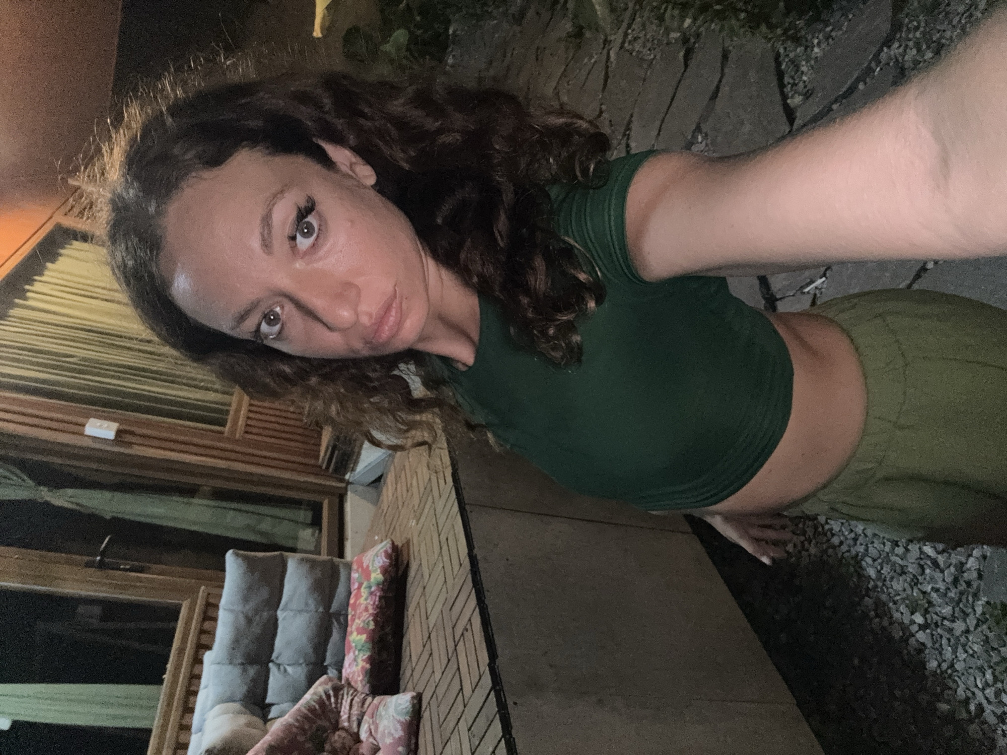 The image shows a woman taking a selfie. She has long, curly dark hair, and she is wearing a short-sleeved green top and light green pants. She is standing outside, likely in a backyard or patio setting. Behind her is a building with a brick facade and a wooden exterior. There is outdoor furniture with cushions visible. The ground is paved with stone and gravel. The lighting is dim. It is not possible to verify if this image was taken in the city of Hà Giang 1 Ward, Vietnam, or to prove its authenticity.