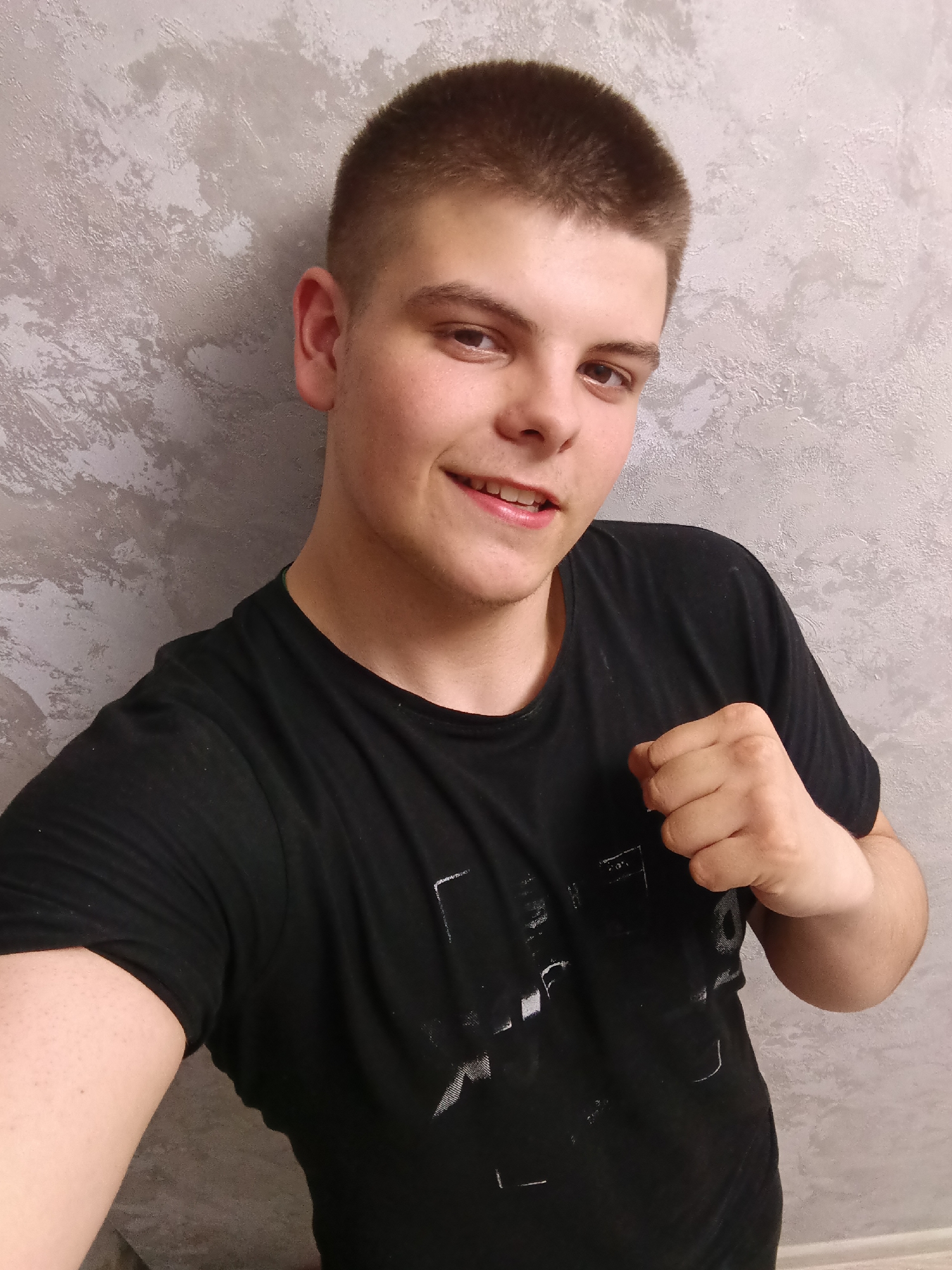 A fair-skinned young man with short, dark hair and brown eyes is shown from the chest up. He wears a black t-shirt and smiles slightly. His right hand is clenched into a fist, held up toward his chest. The background is a mottled gray wall. The location is Unknown, Unknown.