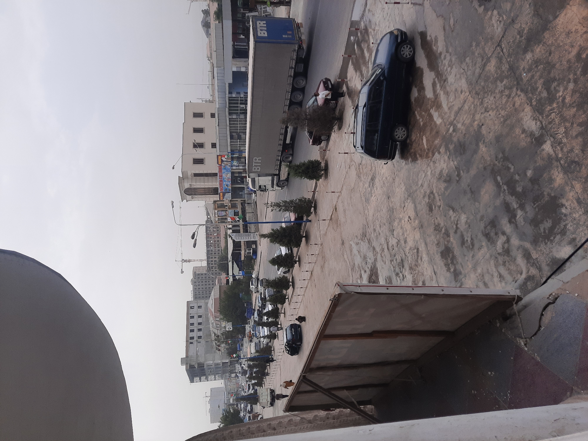 The image depicts a street scene in Hulbuk, Tajikistan, with buildings under construction and a busy road. In the foreground, a large billboard structure is visible on the left, partially obscuring the view. The ground in the foreground appears wet and cracked, with puddles reflecting the overcast sky. A dark-colored station wagon is parked on the wet ground. Further down the street, a large semi-trailer truck with "BTR" branding is driving on the road. Several other cars are also present on the road, and a few people are walking along the sidewalk. Trees line the sidewalk, and various buildings, some under construction with visible scaffolding, form the urban backdrop. The sky is hazy and overcast.