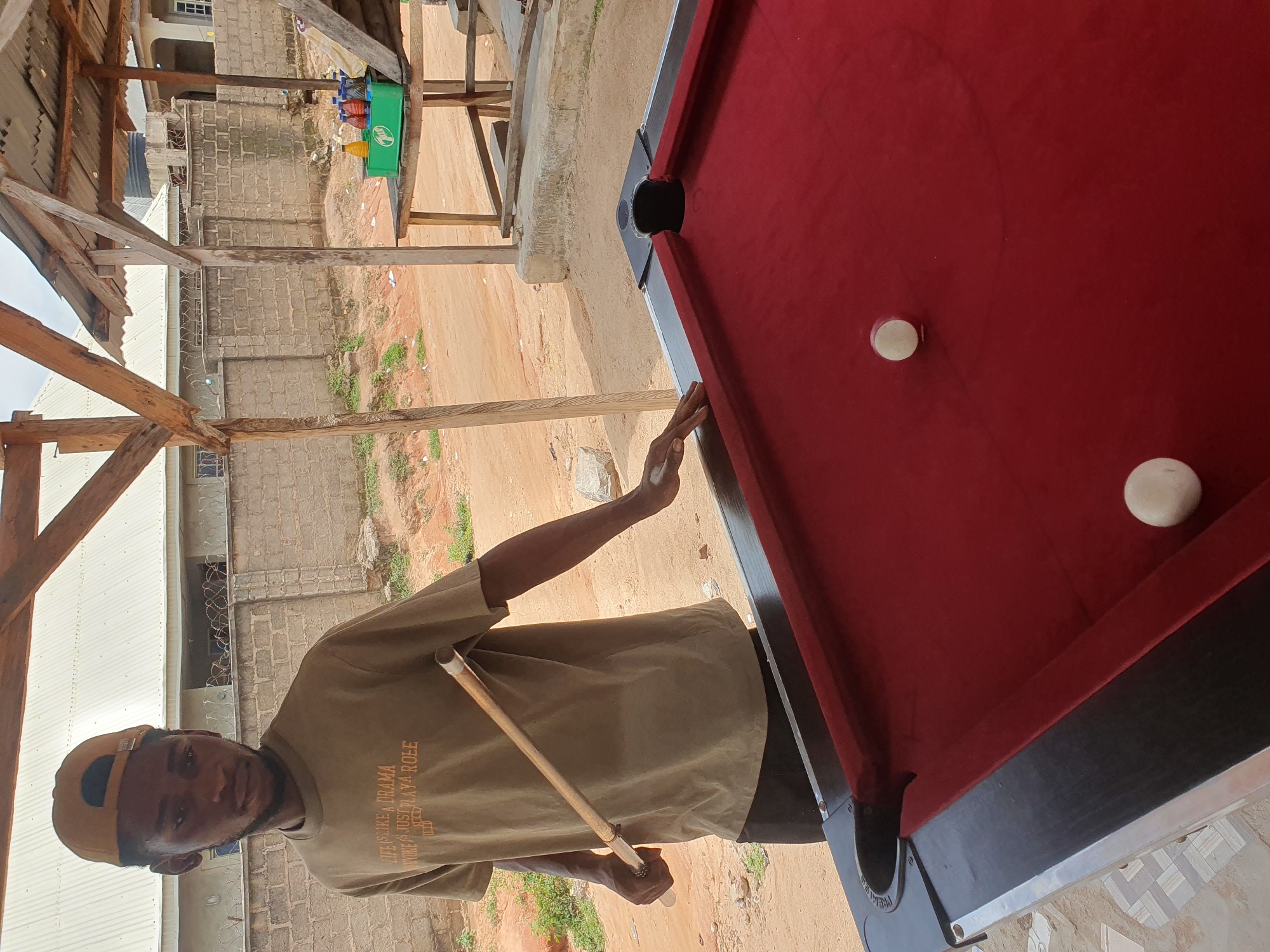 In Lokoja, Nigeria, a young man stands next to a red billiard table, holding a cue stick. He is wearing a light brown t-shirt with the text "LIFE IS LIKE A DRAMA, YOU WORK IT JUST PLAY A ROLE" and a tan baseball cap. The scene is outdoors, under a wooden shelter with a corrugated metal roof. The ground is dirt, and in the background, a brick building and some vegetation are visible. Two white billiard balls rest on the table. The time of day appears to be daytime, with ample natural light. The overall impression is of a casual setting, possibly a local hangout or recreational area.