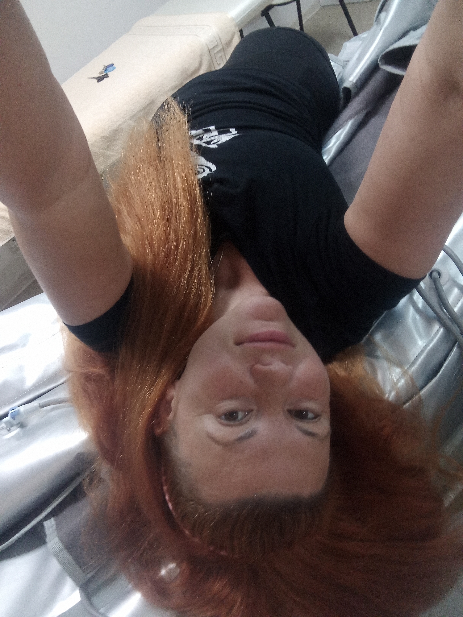 A woman with long, reddish-brown hair is lying on her back, with her head towards the bottom of the frame. She is wearing a black t-shirt and black leggings. Her arms are raised above her head. She appears to be in a room with a light-colored bed or couch in the background and a silver, possibly metallic, surface beneath her. There are some medical tubes or cables visible near her head. The lighting is bright, casting some shadows.