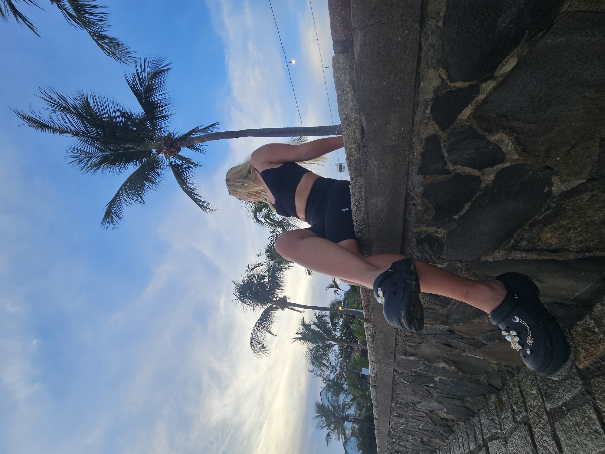 A woman with blonde hair is sitting on a stone wall in Phu Thuy Ward, Vietnam. She is wearing a black sports bra and black shorts. Her legs are crossed and her feet are dangling over the edge of the wall. She is wearing black Crocs with charms. The wall is made of large, dark stones. Behind the woman, there are palm trees and a blue sky with some clouds. There is also a string of lights visible in the distance. The overall scene appears to be at dusk or early evening.