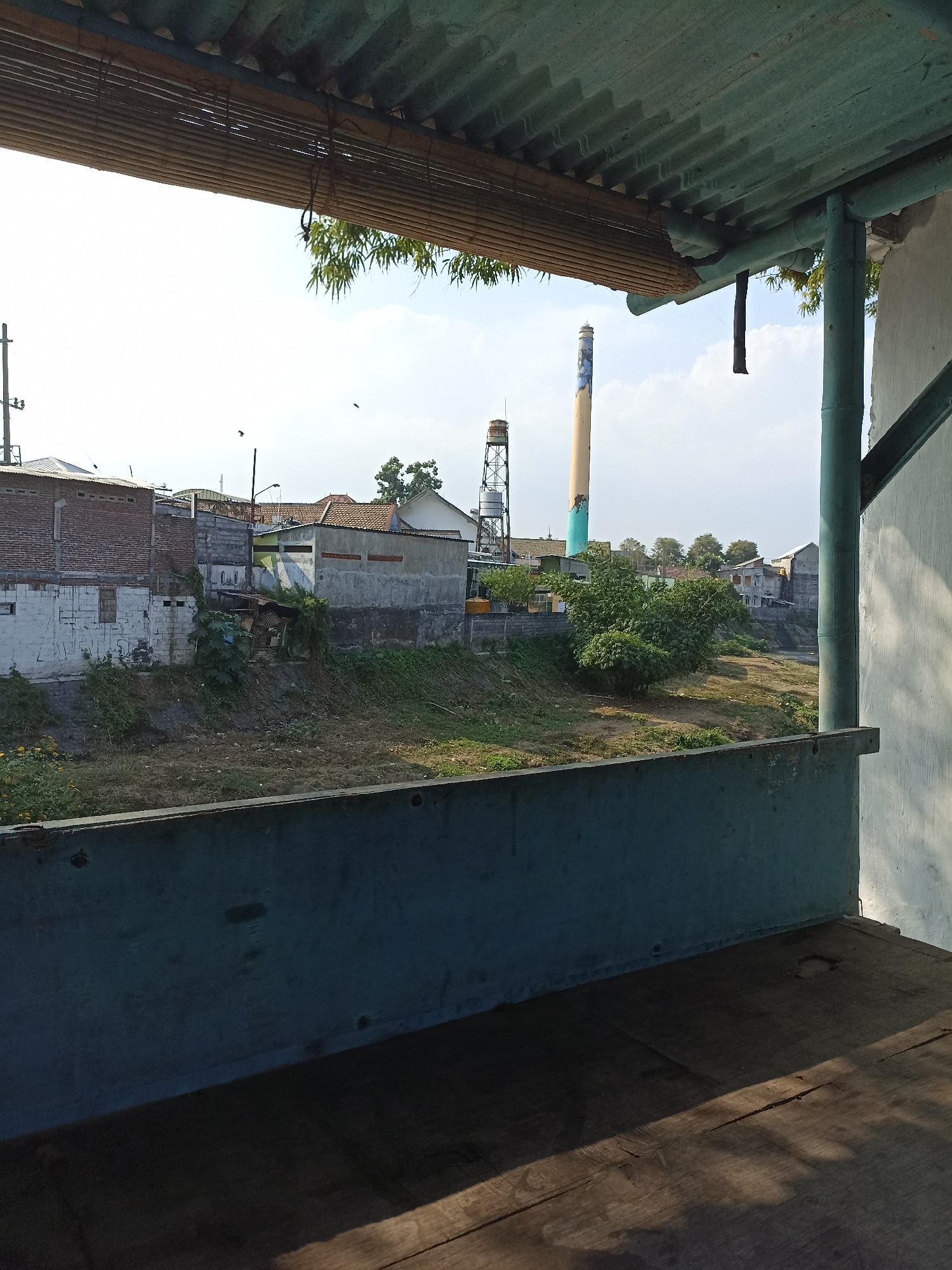 The image captures a view from an elevated wooden platform, looking out over a dry canal bed towards a cityscape under a bright, hazy sky. In the foreground, the dark, weathered wooden planks of the platform are visible, with a blue metal railing along the edge. Beyond the railing, the dry canal is filled with patchy dry grass and some green vegetation.
The middle ground features a collection of buildings. On the left, there's a brick building with white-painted lower sections and a corrugated metal roof. Adjacent to it are more modern structures with concrete walls and tiled roofs. A tall, slender industrial smokestack with blue and yellow bands dominates the right side of the mid-ground. Behind it, a water tower on a metal lattice structure is visible. A cluster of trees provides some greenery amidst the buildings.
In the background, more houses with tiled roofs are scattered, and the sky is bright with scattered clouds. The presence of the smokestack and water tower suggests an industrial area within or on the outskirts of Lumajang, Indonesia. The overall scene conveys a sense of a semi-urban or industrial environment with evidence of arid conditions due to the dry canal. There are no visible people or animals in the frame.