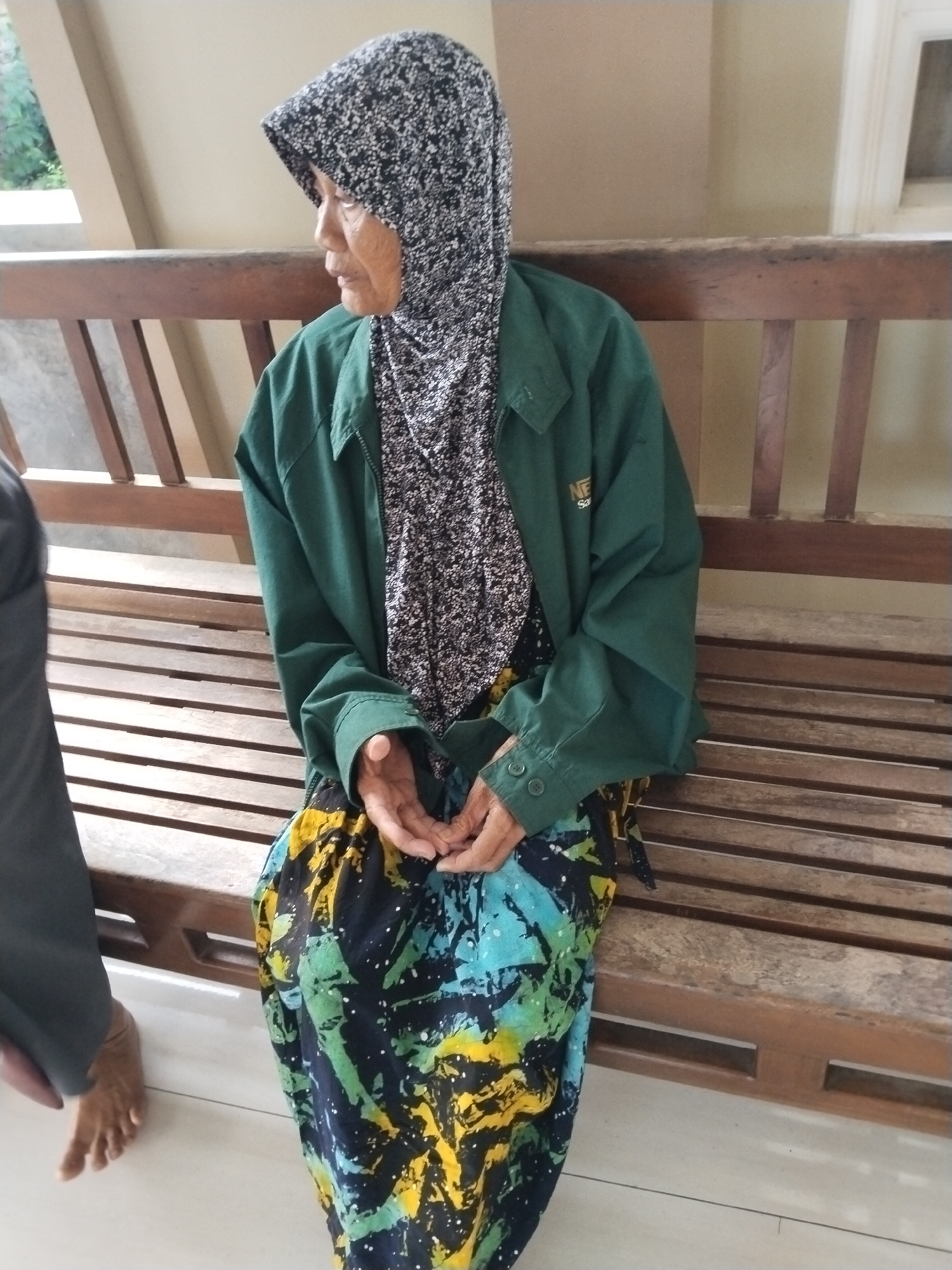 An elderly woman wearing a patterned headscarf and a green jacket over a floral blouse sits on a wooden slatted bench. She is also wearing a colorful batik skirt. Her hands are clasped in her lap. To her left, a person wearing dark pants is partially visible, with one bare foot on the tiled floor. The background consists of a wall and a window. The scene is likely outdoors, possibly in a public waiting area or a tranquil outdoor setting in Karanggandu, Indonesia.