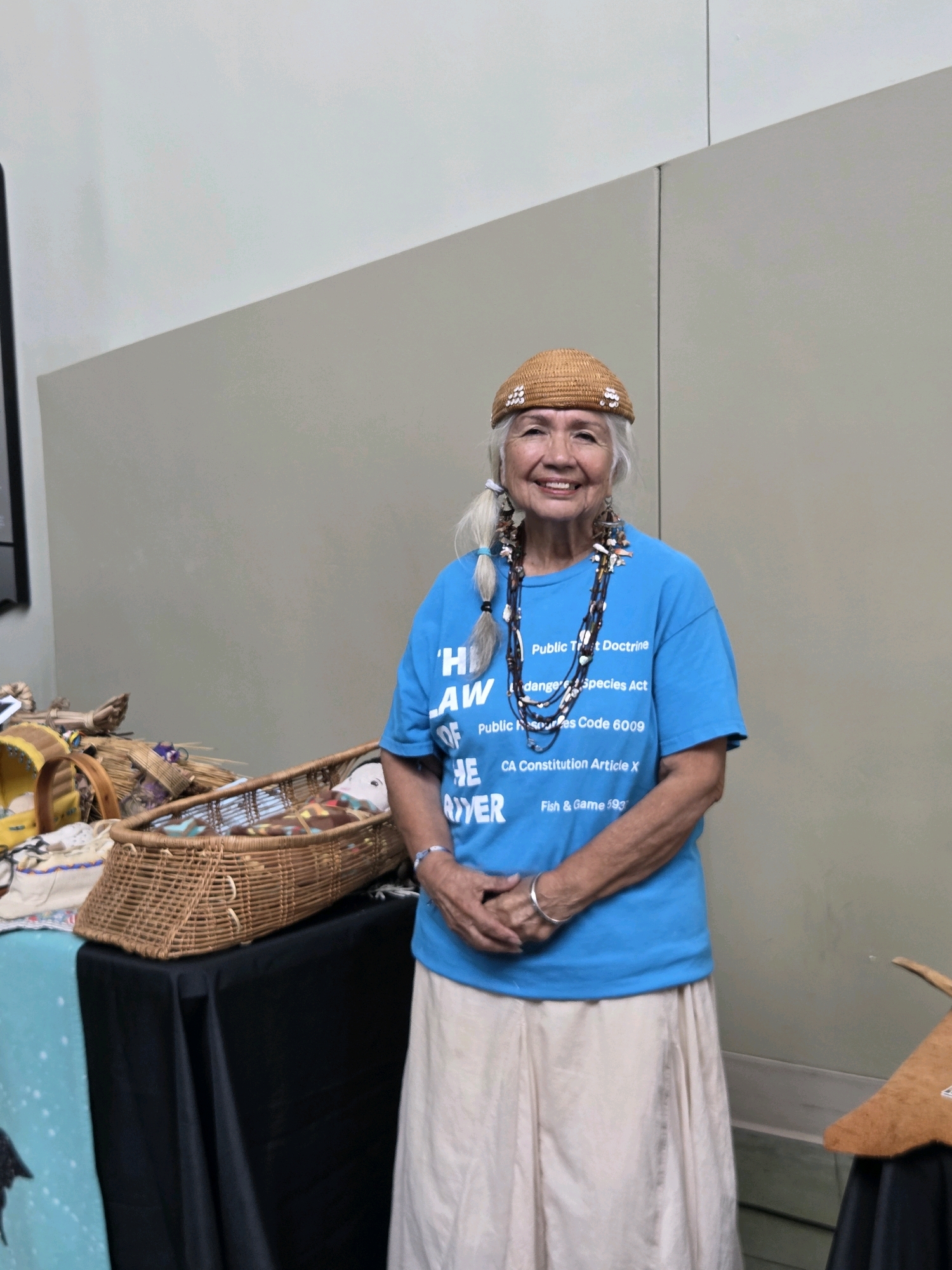A smiling elderly woman of apparent Native American descent stands in front of a wall in what appears to be an indoor community space, possibly a cultural event or exhibition. She is wearing a small, woven, brown hat adorned with small, white, sequin-like objects, and a bright blue t-shirt with white text. The text on her shirt lists various legal codes and acts, including "Public Trust Doctrine", "Endangered Species Act", "Public Resources Code 6009", "CA Constitution Article X", and "Fish & Game 593". She is also wearing multiple strands of beaded necklaces in various colors and materials, and large, decorative earrings. Her arms are crossed, and she has on a simple silver bracelet. A long, light-colored, pleated skirt is visible below the t-shirt. To the left of the woman, a table is covered with black and teal-colored cloths. On the table, there are various woven baskets and what appear to be craft materials or artifacts. One prominent basket is large and oval-shaped, containing an assortment of colorful items. To the right of the woman, another table is partially visible, covered by a black cloth, with a brown, horn-like object on display. The wall behind her is a neutral, two-toned color, with a distinct horizontal line dividing the upper and lower sections. The lighting suggests an indoor environment with artificial light. The scene is static, with no apparent action or interaction depicted other than the woman's posed stance.