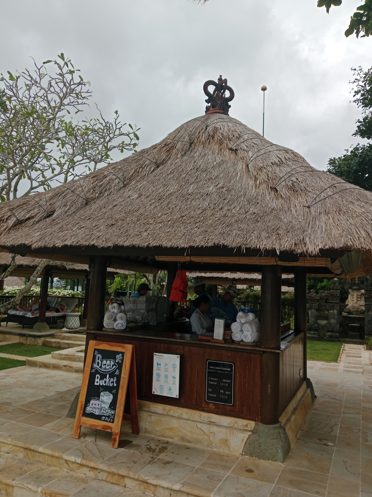 This image shows a thatched-roof bar at a resort in Kuta Selatan, Indonesia. The bar has a wooden counter and is staffed by at least three people. A chalkboard sign in front of the bar advertises "Beer Bucket" with a drawing of a beer. There are also rolled towels stacked on the counter and a life jacket hanging to the right. In the background, guests are seen relaxing on lounge chairs. The resort appears to be in a tropical setting with lush greenery and stone structures.