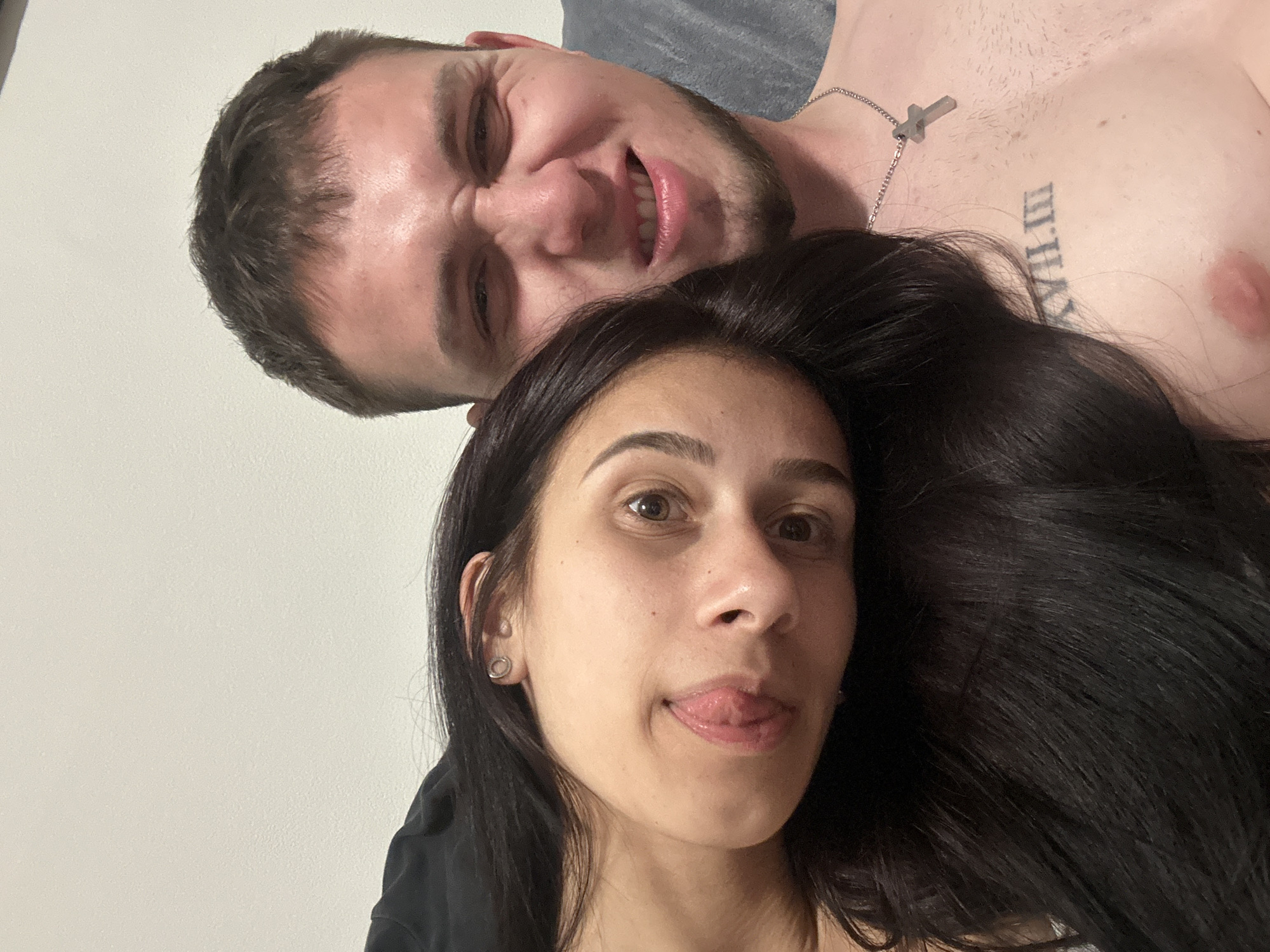 The image shows two individuals in a close, selfie-style composition. A woman with long, dark hair is in the foreground, sticking her tongue out. A man is positioned behind and above her, making a face with his mouth open and eyes narrowed. He has short brown hair and a beard. The man is shirtless and wearing a silver cross necklace. A tattoo in Roman numerals is visible on his upper chest. The background includes a plain white wall on the left and what appears to be a grey-toned blanket or surface on the right. There is no contextual information available to pinpoint the location as Plovdiv, Bulgaria.