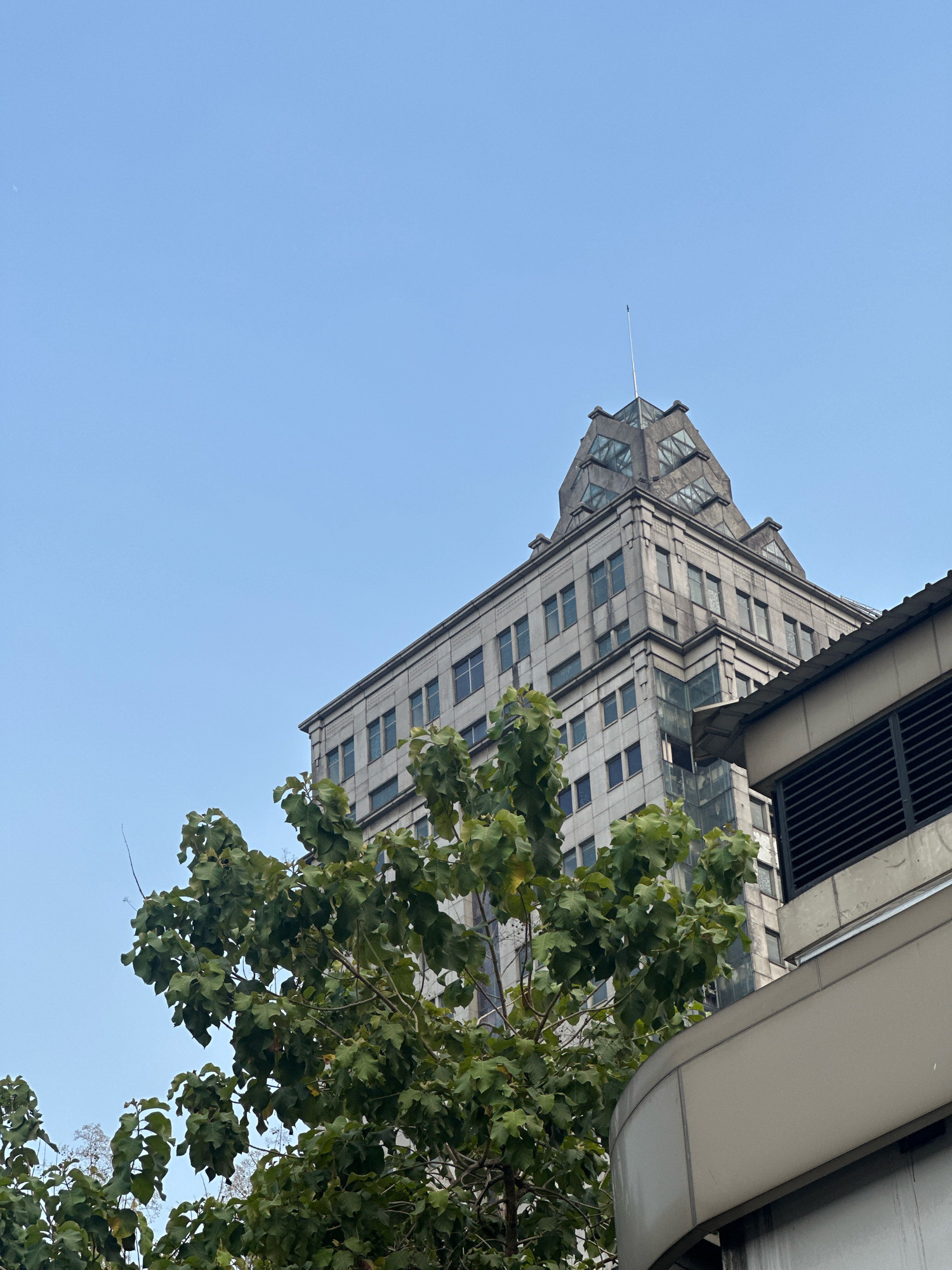 The image shows a grey, multi-story building with a distinctive stepped, pyramid-like top, and a slender flagpole. The building's facade is composed of rectangular panels and numerous windows. In the foreground, a large tree with lush green leaves partially obscures the lower levels of the building. To the right, a portion of another structure with horizontal louvers is visible. The sky is a clear, pale blue. This scene is likely located in South Jakarta, Indonesia, given the architectural style of the building and the type of vegetation. There are no people or animals visible in the image.
