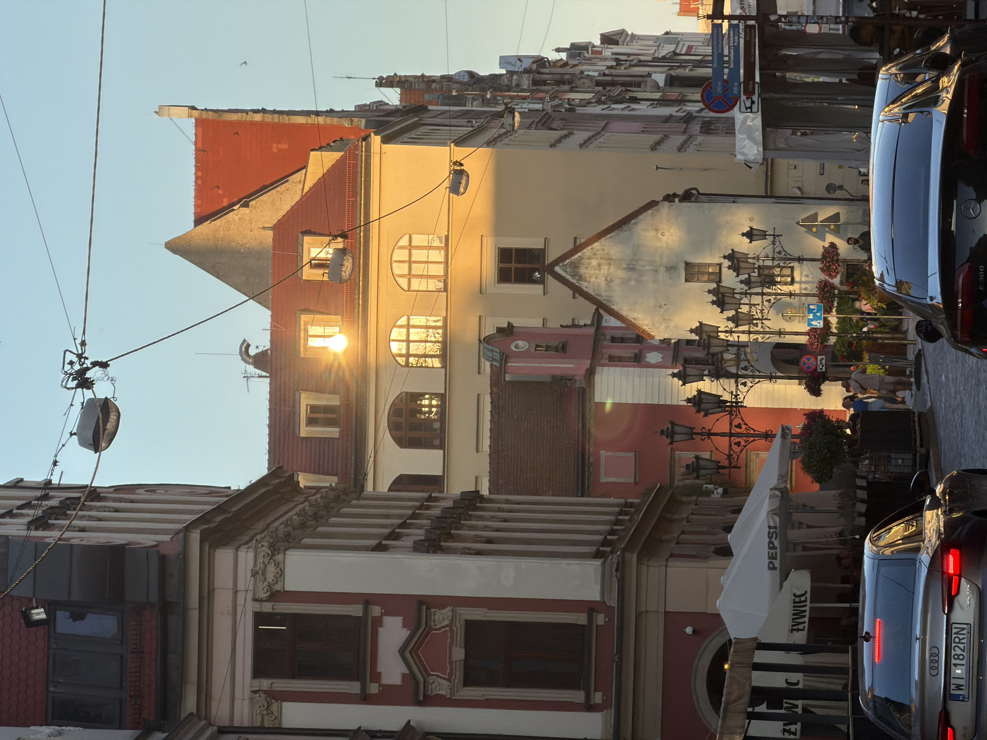 The image depicts a street scene in Wrocław, Poland, with traditional European architecture. The sunlight casts a warm glow on the buildings, highlighting their ornate facades and red-tiled roofs. On the left, a building with a red and white facade features classical architectural elements like columns and decorative moldings around the windows. Across the street, a row of buildings displays varying architectural styles, some with arched windows and steeply pitched roofs.
In the foreground, the rear of a silver Audi sedan with license plate "WI 182RN" is visible, its brake lights illuminated. Further down the street, a black Mercedes-Benz sedan, identified by "E300" on its trunk, is also present.
Several people are visible walking on the sidewalk and in the street. Some are seated under umbrellas belonging to an outdoor cafe. A street lamp post with multiple lights stands prominently, decorated with hanging flower baskets. Signage is present, including a blue directional sign with Polish text and a red and blue circular traffic sign. The street appears to be paved with cobblestones. Overhead, electrical wires and street lamps are strung across the street, common in older urban areas. The sky is clear and blue, suggesting it is daytime, likely late afternoon or early evening due to the warm sunlight.