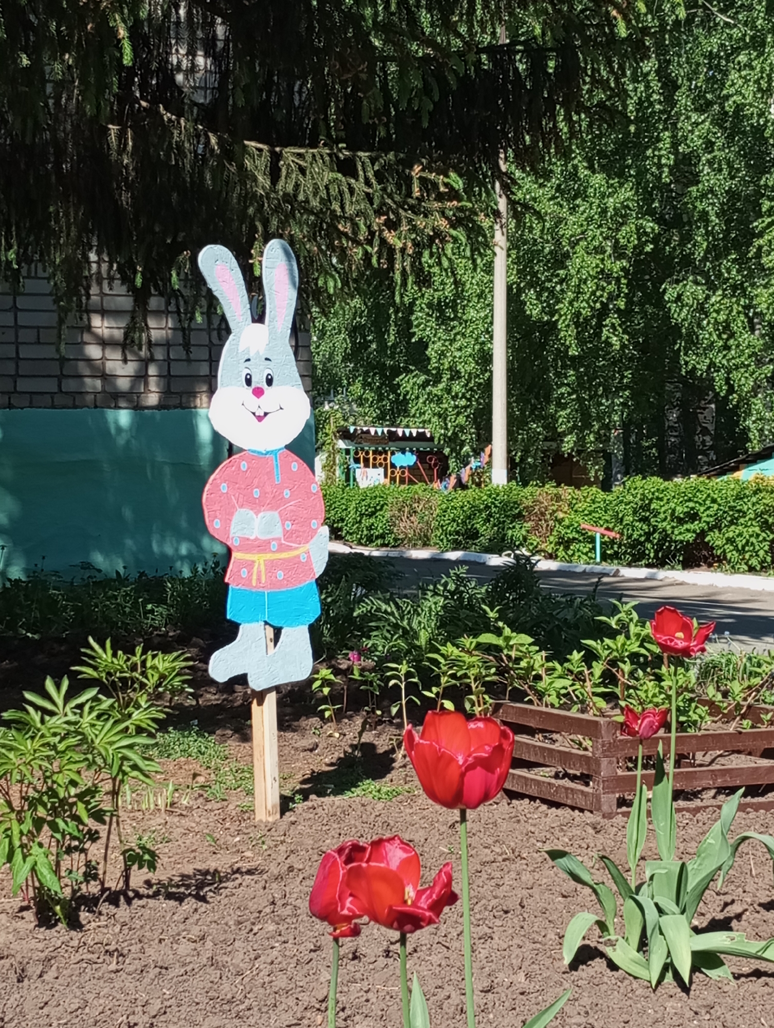 The image shows a garden setting in Izhevsk, Russia, on a sunny day. In the foreground, three vibrant red tulips are in bloom, planted in dark brown soil. Behind the tulips is a wooden fence or border. To the left, there's a painted plywood cutout of a rabbit wearing a red shirt and blue pants, placed in the garden as a decorative element. In the background, a building with a light brick facade and teal paneling is visible on the left. Further back, there are lush green bushes and trees, with what appears to be a small, colorful structure, possibly a children's playhouse, in the distance. A utility pole is also visible in the background. The scene appears to be a public garden or park area within the city.