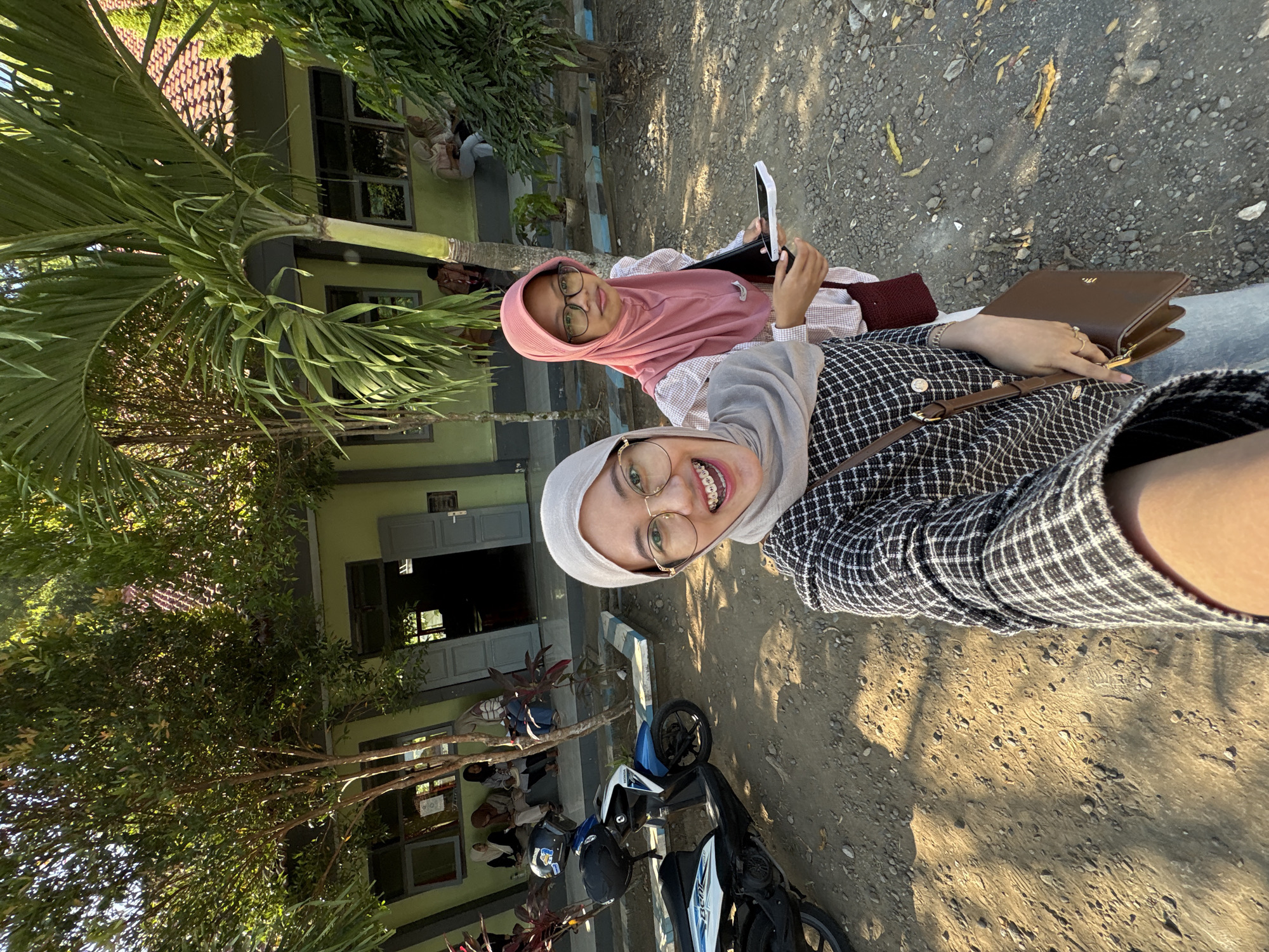 In Gunungsari, Indonesia, two young women are captured in a selfie outside a school building. The woman in the foreground, wearing a grey hijab and a checkered top, smiles broadly at the camera, her braces visible. She holds a brown shoulder bag. To her right, another woman in a pink hijab and glasses looks towards the camera, holding a notebook and a phone. They are standing on a dusty ground next to a blue and white scooter. In the background, other individuals are seated on a raised platform in front of the school, which has a green facade and a red tiled roof. Lush green trees and palm fronds provide shade, suggesting a warm, sunny day.