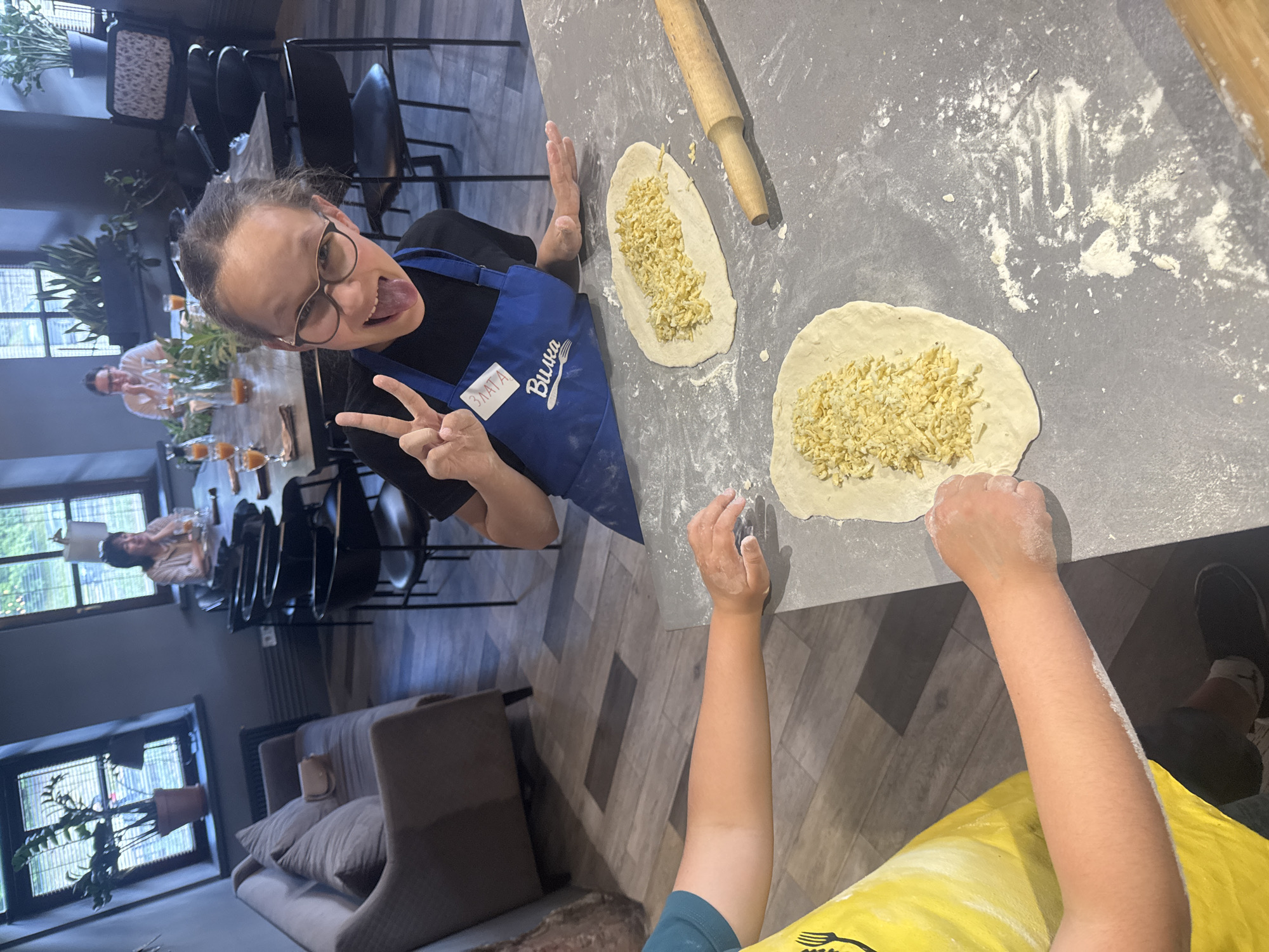 The image captures an engaging indoor cooking session, likely a masterclass or a casual gathering, with a vibrant, playful atmosphere. The scene is shot from a high-angle, slightly tilted perspective, focusing on the food preparation area.
The main subject is a young person, identified as Zlata by her name tag, wearing glasses and a blue apron over a black top. She looks directly at the camera with a cheerful, humorous expression, sticking out her tongue and making a peace sign with her left hand. Her right hand is visible, dusted with flour. She is positioned next to a gray countertop heavily dusted with flour, where two pieces of flattened dough are laid out, each generously filled with shredded cheese or a similar light-colored ingredient. A wooden rolling pin rests nearby on the counter.
Another person's arm, wearing a yellow top or apron, is visible in the foreground, also engaged in the food preparation, indicating a collaborative activity. In the background, two other individuals are seated at a large wooden table, enjoying drinks served in tall glasses. Potted plants adorn the table and windowsills, adding a touch of greenery to the modern, somewhat industrial-chic interior. Large windows suggest natural light, indicating it is daytime. The room features dark walls, patterned wooden flooring, and a mix of seating including black chairs and comfortable sofas in the distance. The text "ЗЛАТА" and "Вилка" (meaning "Fork," accompanied by a fork icon) are clearly visible on the blue apron, confirming Russian language and aligning with the given context of Izhevsk, Russia.