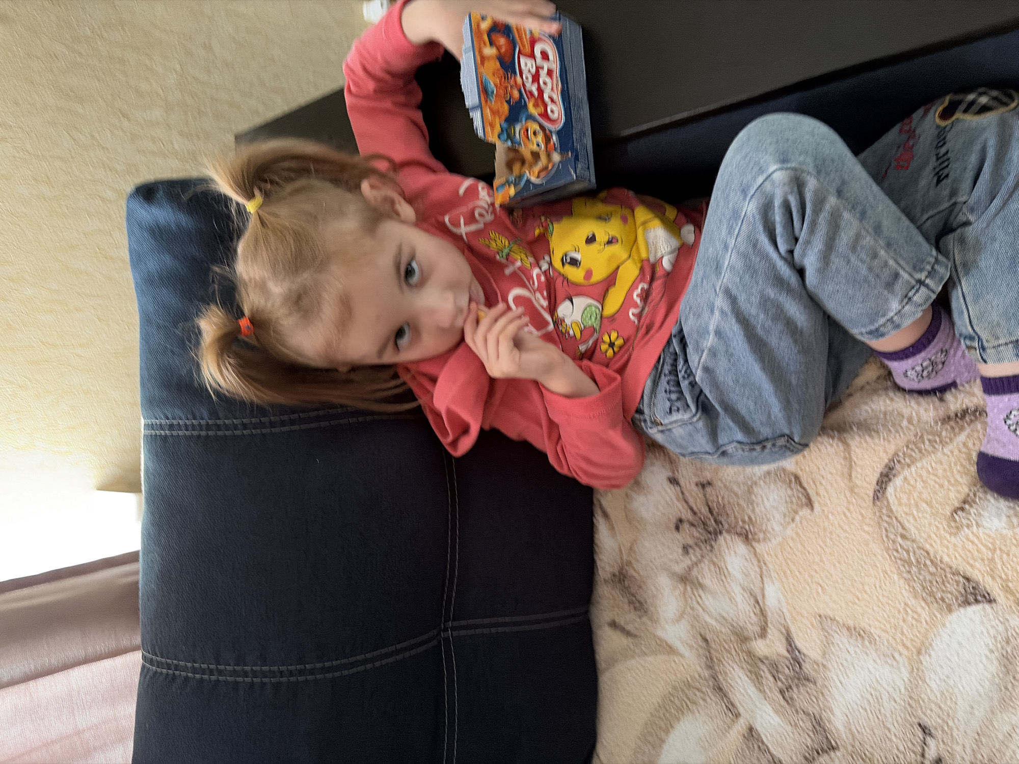 A young girl with blonde hair pulled into two pigtails is sitting on a dark blue couch with a patterned blanket. She is wearing a pink sweatshirt with a yellow cartoon character on it and blue jeans. The girl is holding a small snack box with colorful cartoon characters and appears to be eating a snack from it. She is looking at the camera with a neutral expression. The background shows a wall and a curtain. The scene is indoors, likely in a residential setting. There is no visual information to confirm a specific location in Tambov, Russia.