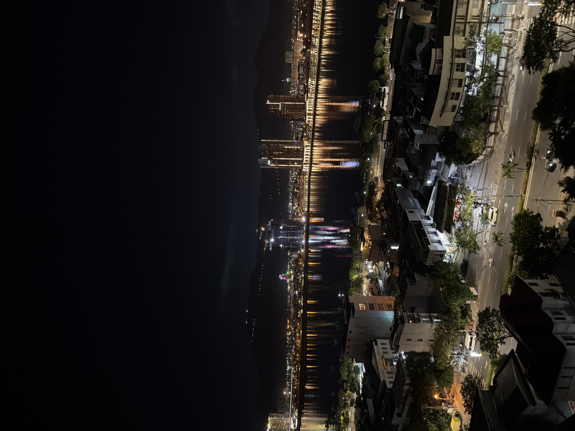 This is a night aerial view of the Han River in Da Nang, Vietnam. The river reflects the city lights from buildings and streetlights on both banks. In the background, mountains are visible with scattered lights. On the foreground bank, there are residential buildings and streets with some vehicles and trees. On the far bank, a skyline of illuminated modern buildings and a construction site is visible.