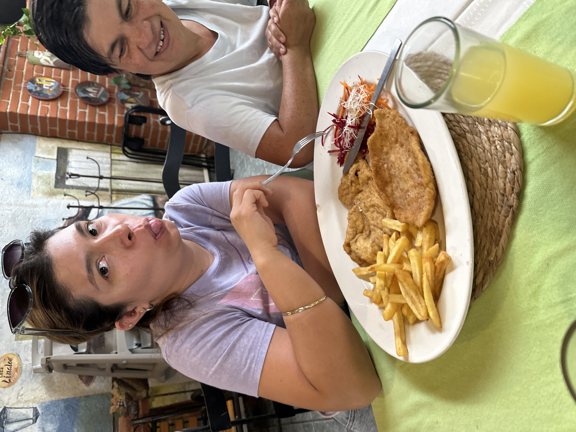 Two women are seated at a table in a restaurant. A plate of food with fried meat, fries, and shredded carrots/beets is on the table, along with a glass of yellow liquid. One woman is smiling at the other woman, who has stuck out her tongue. The restaurant has brick accents and decorative plates on the wall. The woman sticking her tongue out is wearing sunglasses. A sign with the name "Lindloe" is also visible in the background. Based on the background and the style of restaurant, this image was taken in Puebla, Mexico.
