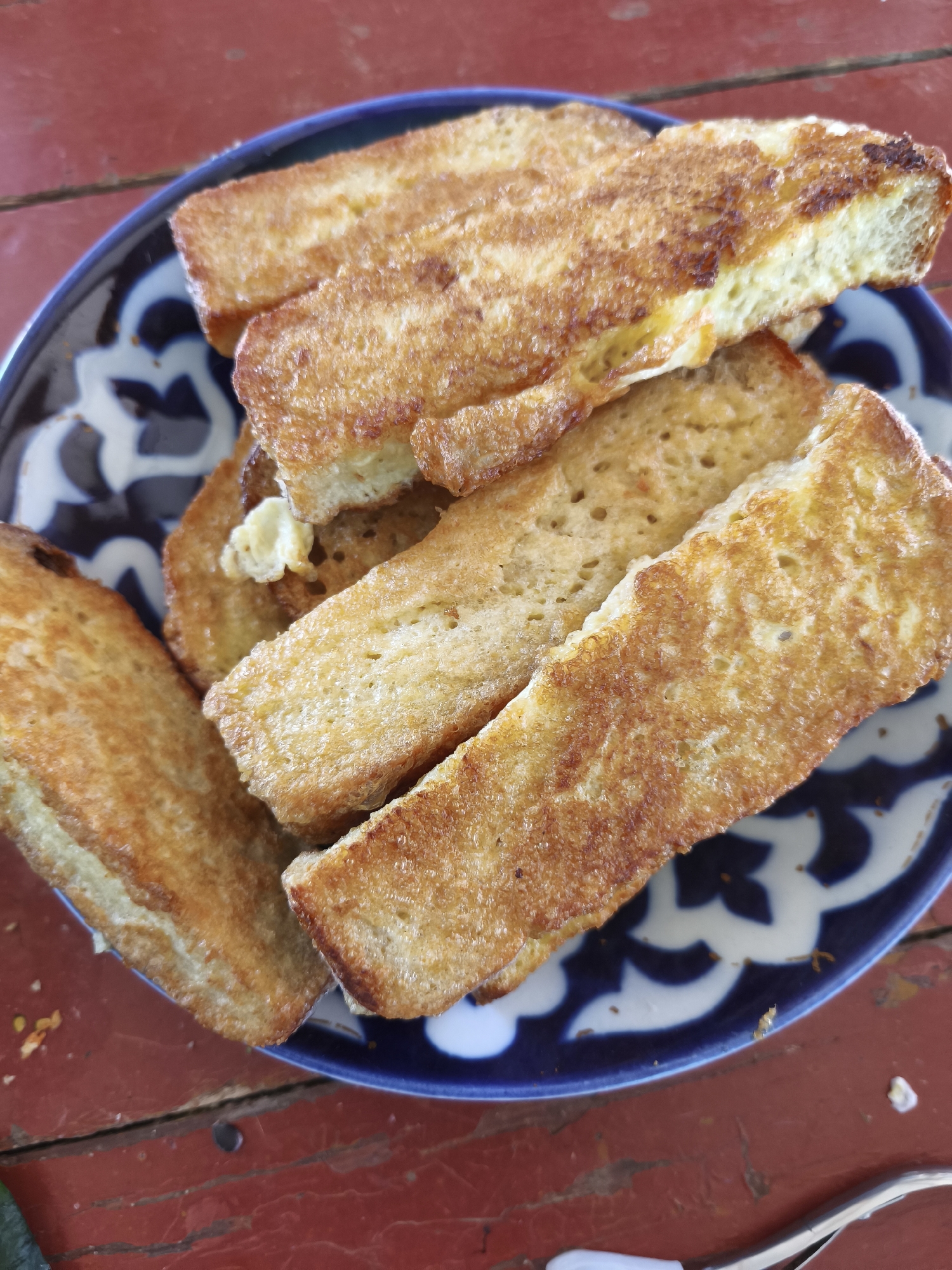A plate of French toast sits on a red wooden table. The French toast is golden brown and appears to be cut into strips. The plate is blue and white with a decorative pattern. The table is made of planks and has a rustic appearance. The scene is outdoors, as indicated by the lighting and the weathered appearance of the table. The location is Ленинское сельское поселение, Ukraine.
