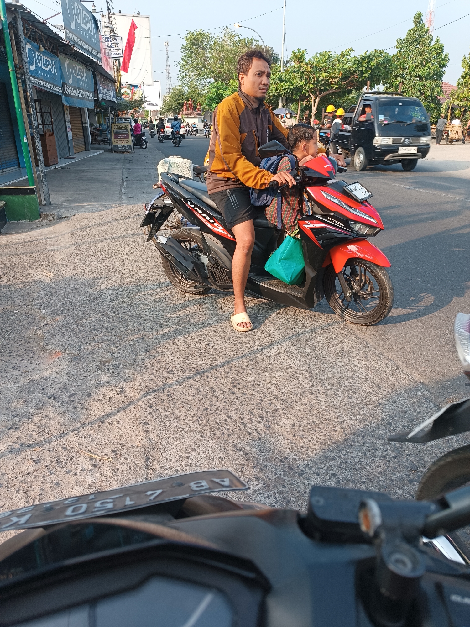 In Imogiri, Indonesia, a man in a brown and orange jacket and black shorts rides a red and black Honda Vario motorcycle. A young boy, wearing a patterned shirt, sits in front of him, also on the motorcycle. The man is looking to the left, while the boy is looking forward. A bright green bag is hanging from the handlebars. The motorcycle is parked on a paved street, with shadows from trees and buildings cast across the ground. In the background, other motorcycles and a black car are visible, along with shops and trees lining the street. The sun is low in the sky, indicating it is likely late afternoon or early morning.