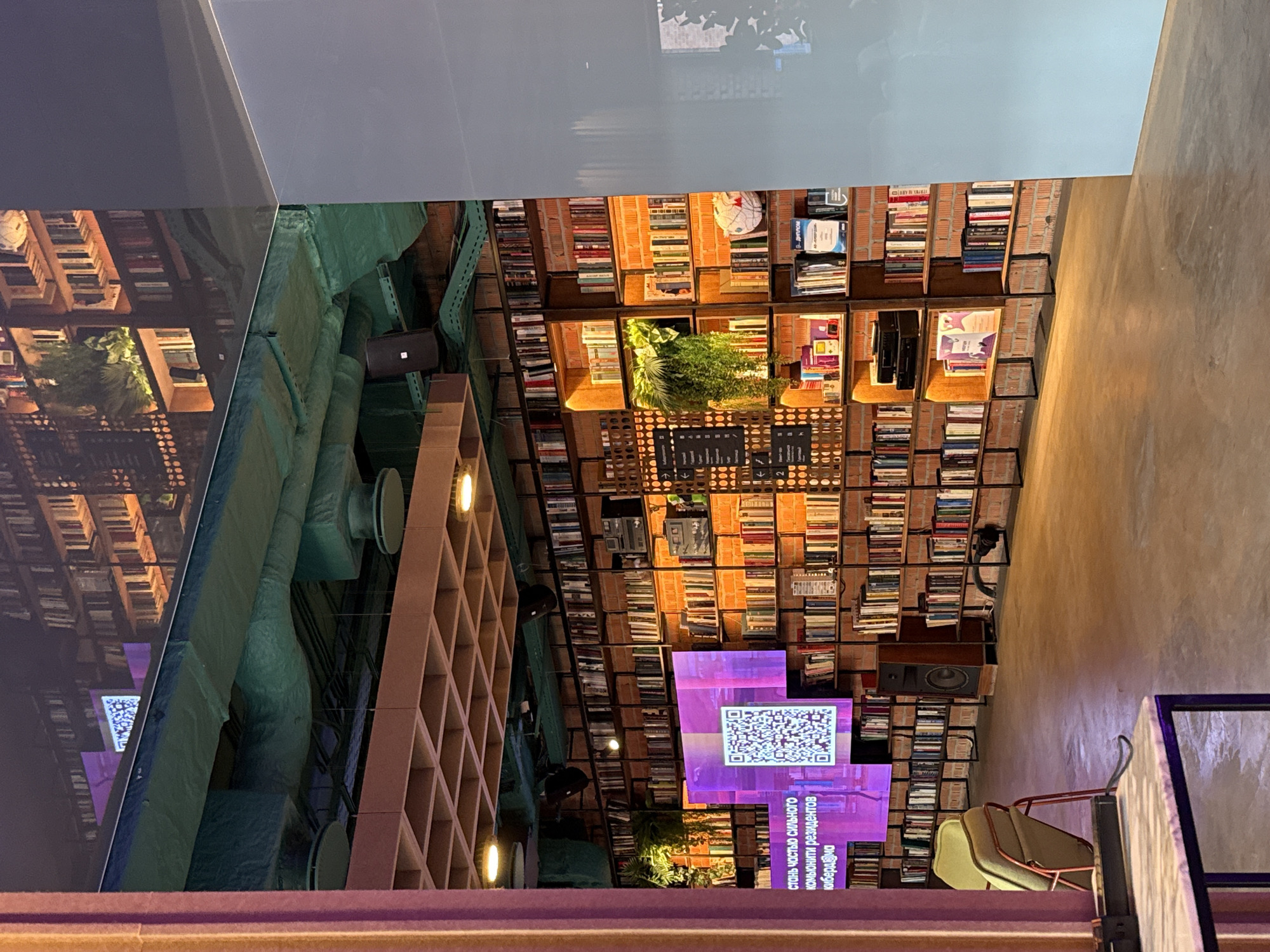 The image displays the interior of a contemporary space, likely a library, bookstore, or a modern cafe/co-working area, located in Moscow, Russia. The primary feature is a large wall of bookshelves, extending from floor to ceiling, filled with numerous books. These shelves are interspersed with decorative green plants and a white globe. The wall behind the shelves appears to be made of brick.
On the left side of the room, industrial-style green-wrapped ventilation ducts run across the ceiling. Below these, a light-colored wooden lattice structure is visible, potentially serving as shelving or a room divider. The ceiling above this area is a smooth, reflective surface.
Several digital screens are integrated into the bookshelves. One prominent purple screen displays a QR code and Russian text below it. The text reads: "СТАНЬ ЧАСТЬЮ СИЛЬНОГО СООБЩЕСТВА РЕЗИДЕНТОВ КИБЕРД@ЧА," which translates to "BECOME PART OF A STRONG COMMUNITY OF CYBERD@CHA RESIDENTS." "КИБЕРД@ЧА" (Cyberdacha) is a distinct name, likely referring to a tech or creative community. Another screen appears to show a world map. A large black speaker is also positioned on one of the shelves.
In the foreground on the right, a section of a table and two upholstered chairs are visible, indicating a seating area. The lighting in the room is warm and ambient, creating an inviting and cozy atmosphere. No people are present, and no specific activity is taking place in this static shot. The scene suggests a modern, intellectual, and community-focused environment.