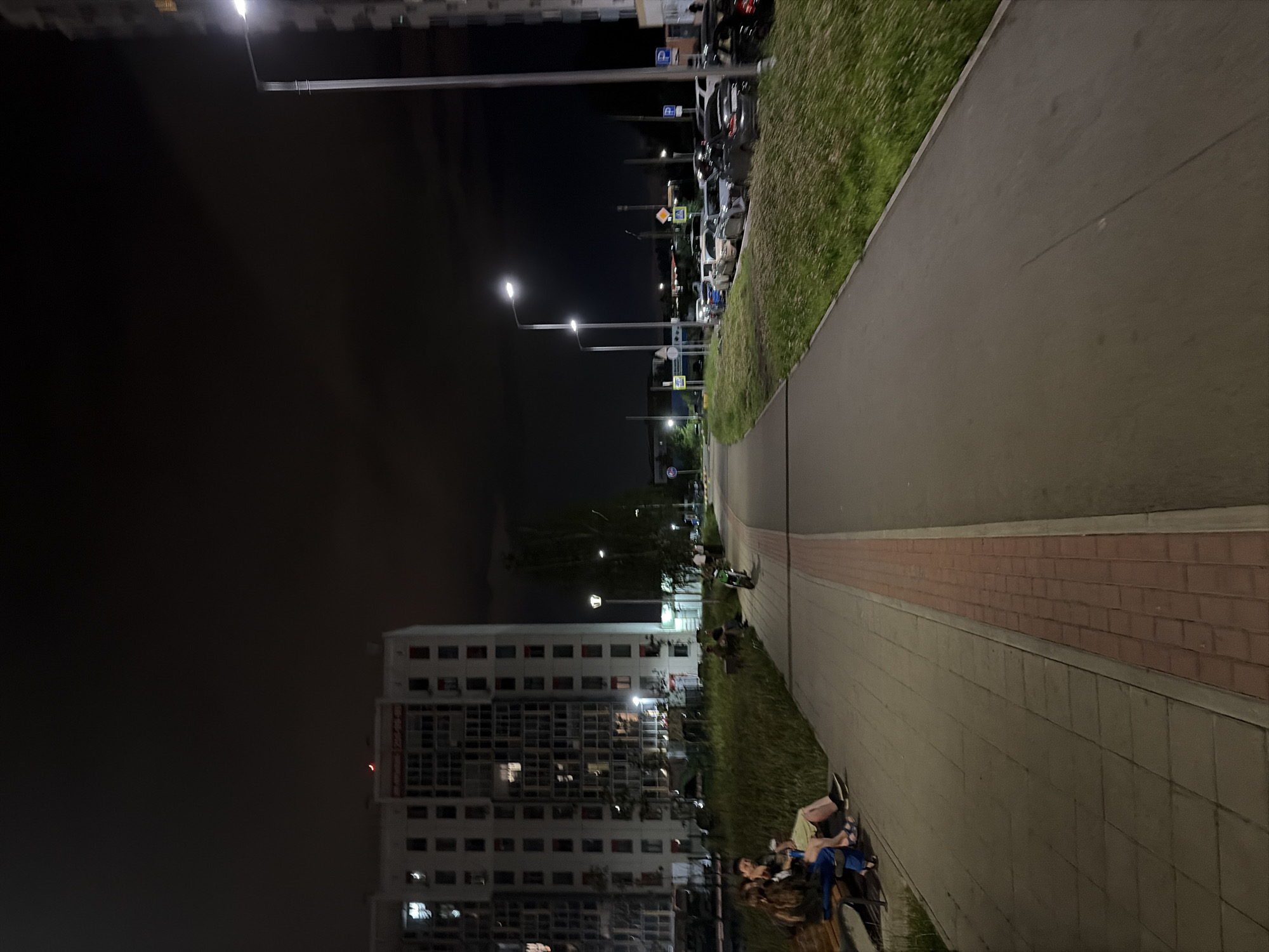 The image depicts an outdoor scene at night, likely in a residential urban area of Yekaterinburg, Russia, characterized by a tall apartment building on the left and a row of parked cars on the right. A paved walkway with a red brick accent runs through the foreground.
On the left side of the walkway, a group of individuals are seated on a bench, appearing to be engaged in conversation. Behind them is a grassy area and the large, multi-story apartment building. The building's windows are illuminated, suggesting occupancy. Streetlights are visible, casting light on the walkway and surrounding areas.
To the right of the walkway, a line of cars is parked along the curb. The sky is dark, indicating nighttime, and clouds are present. Further in the distance, other buildings and streetlights are visible. A lone electric scooter is parked on the walkway in the mid-ground. The overall atmosphere is that of a quiet evening in a city neighborhood.