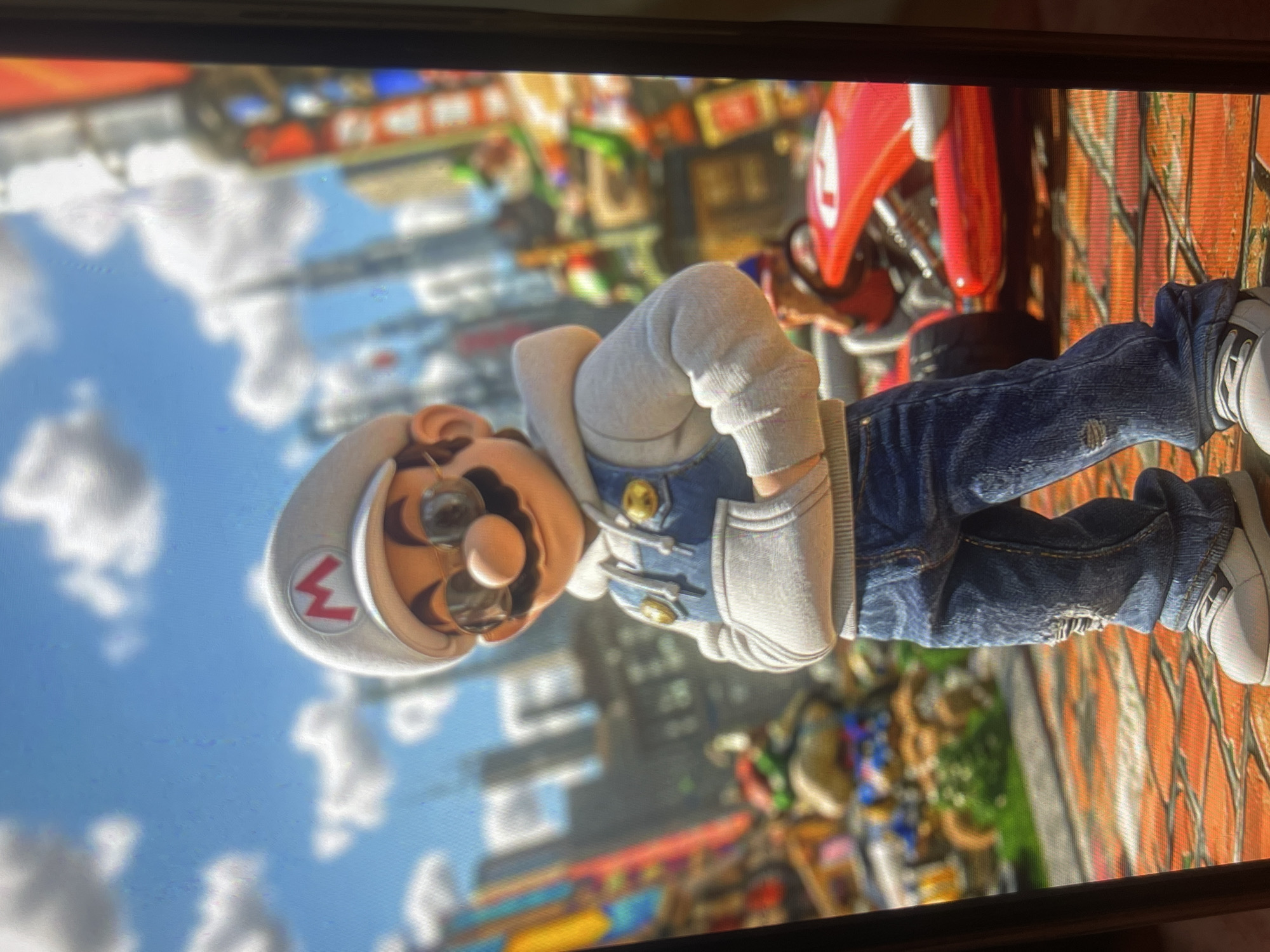 The image shows a character resembling Mario from the Super Mario franchise, dressed in a modern, casual outfit. The character is wearing a white cap with the iconic red 'M' logo, sunglasses, a light gray hooded jacket over denim overalls, and distressed blue jeans with sneakers. The character stands with hands in the pockets of the overalls, looking forward. The background depicts a stylized city scene with tall buildings and vibrant colors, suggestive of a video game environment. A red go-kart is visible to the right of the character. The image does not depict any location in Bishkek City, Kyrgyzstan.