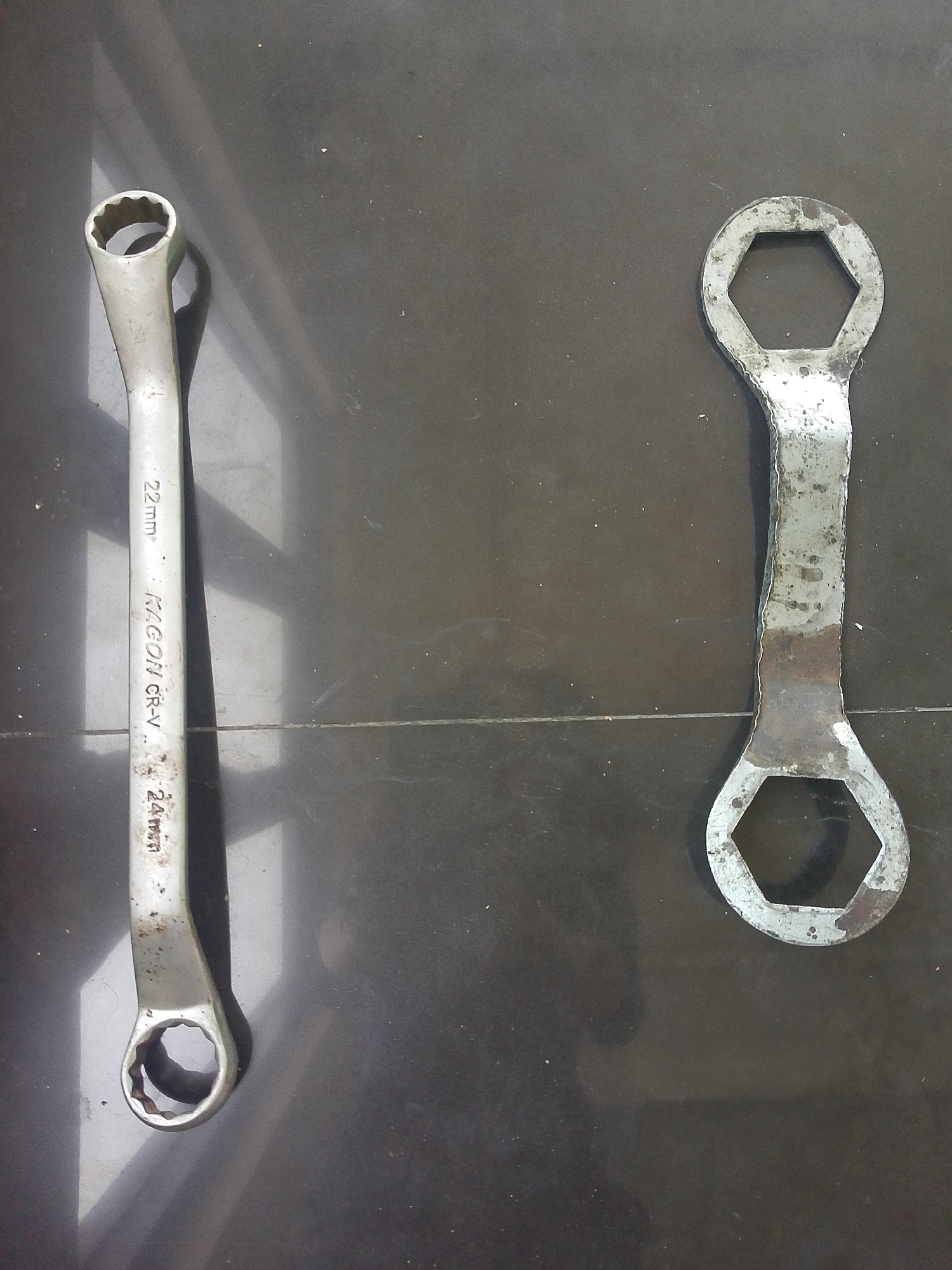 Two wrenches are laid out on a dark, tiled floor. The wrench on the left is longer and appears to be made of polished metal, with markings "22mm", "KAGON CR-V", and "24mm" visible along its shaft. It has a double-ended design, with a 12-point socket on one end and a 6-point socket on the other. The wrench on the right is shorter and appears to be made of a rougher, possibly older, metal with some rust. It also has a double-ended design, with two hexagonal openings of different sizes. The lighting suggests the photo was taken indoors, likely in a workshop or garage, and not in Situwangi, Indonesia, as the environment does not reflect the typical characteristics of that city.