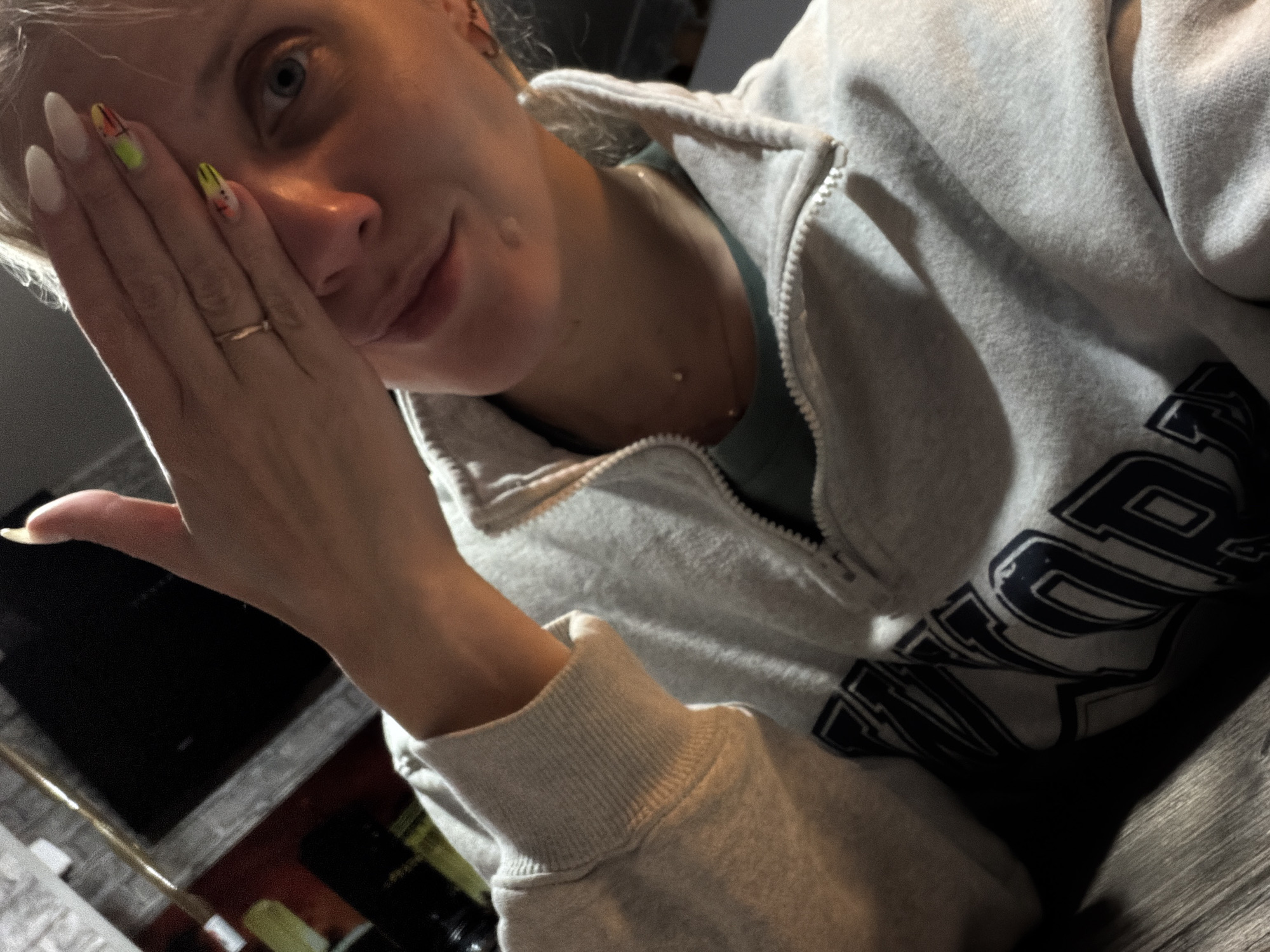 A medium shot shows a fair-skinned person partially obscuring their face with their hand, featuring almond-shaped nails with colorful designs on two of them, while wearing a ring on the ring finger. They wear a partially zipped, light grey sweatshirt with the visible part of a dark green shirt beneath. The sweatshirt features a large, dark graphic design on the right side. The person's face is angled, with a slight smile and light blue eyes. A small mole is visible on their right cheek, and a gold necklace rests on their collarbone. The background shows a dim room with a TV screen and what appears to be various objects or containers.