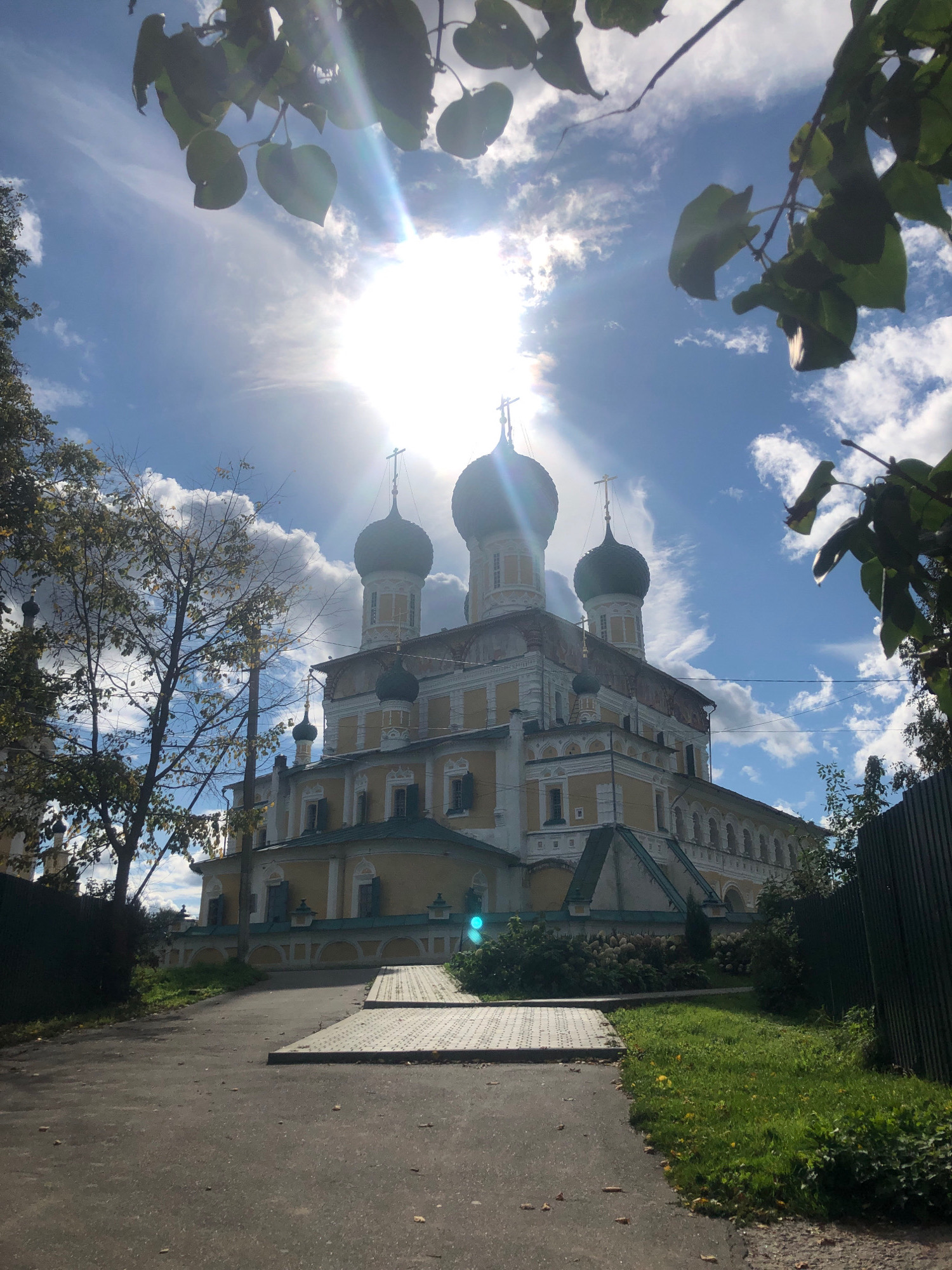 In Tutaev, Russia, a bright, sunny day illuminates a grand yellow and white church with five onion domes, each topped with a cross. The church, adorned with intricate architectural details, stands majestically against a vibrant blue sky dotted with fluffy white clouds. Sunlight streams through the leaves of trees in the foreground, casting a soft glow on the scene. A paved pathway leads up to the church, bordered by lush green grass and bushes. The overall atmosphere is one of serenity and spiritual grandeur, capturing the timeless beauty of this sacred landmark.