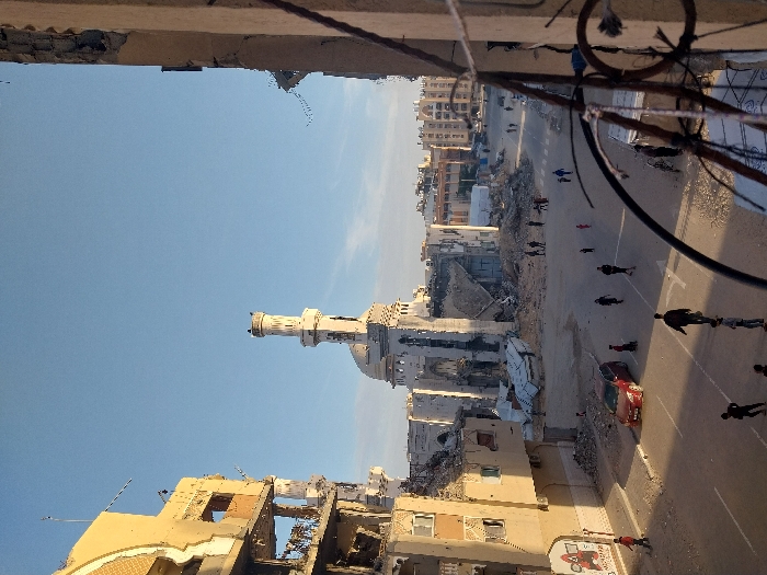 The image presents a severely damaged urban landscape in Khan Yunis, Palestinian Territory, under a clear blue sky. A large, light-colored mosque, featuring a prominent, relatively intact minaret and a dome, stands centrally but shows extensive structural damage to its main body and surrounding structures, with significant sections appearing collapsed or heavily compromised. On both the left and right sides of the frame, multi-story buildings exhibit widespread structural failure, including missing walls, exposed interiors, and shattered windows. Large piles of rubble, concrete debris, and twisted metal are scattered across the ground, particularly in the central area surrounding the mosque.
A paved street, partially covered in dust and debris, runs through the lower portion of the image. Approximately two dozen individuals, including adults and children, are observed walking casually on the street and amidst the rubble. One red four-door sedan is parked or abandoned on the left side of the street. In the immediate foreground on the right, a rusty metal railing and a section of a damaged building are visible, suggesting the photograph was taken from an elevated position within a compromised structure. The scene is illuminated by natural daylight, casting moderate shadows.