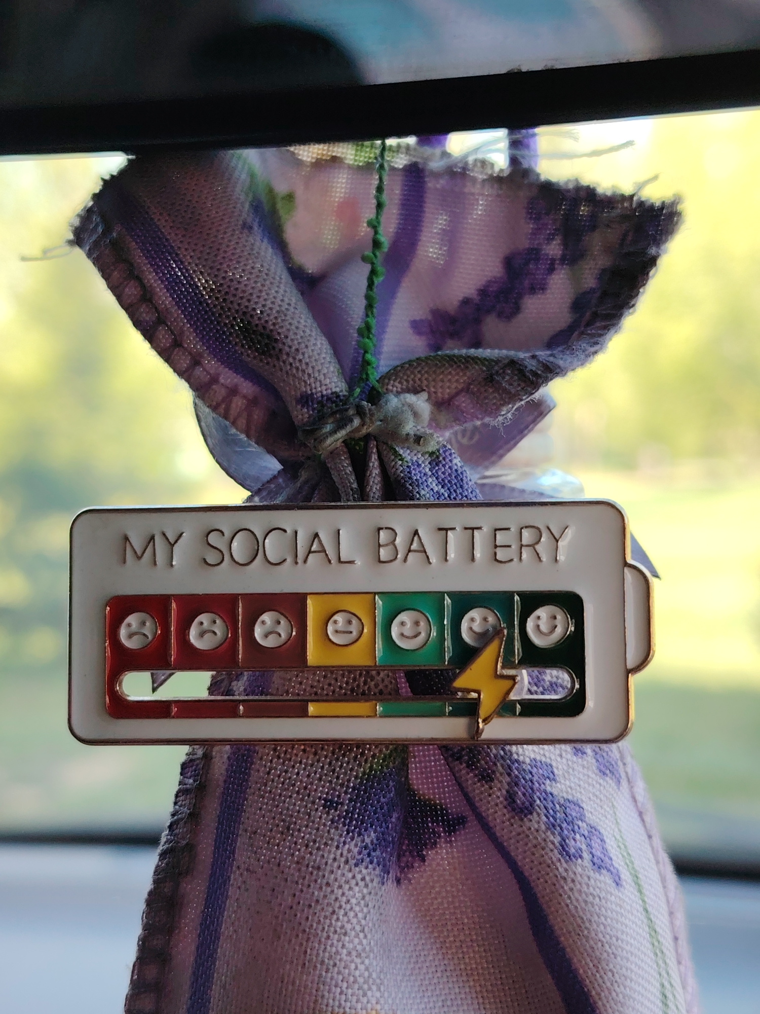 The image shows a "MY SOCIAL BATTERY" indicator hanging from a lavender-patterned cloth, likely suspended from a car's rearview mirror. The indicator shows a progression of faces from sad to happy, indicating the level of social battery. The battery is nearly full, with a yellow lightning bolt indicating the current level. The location is unknown.