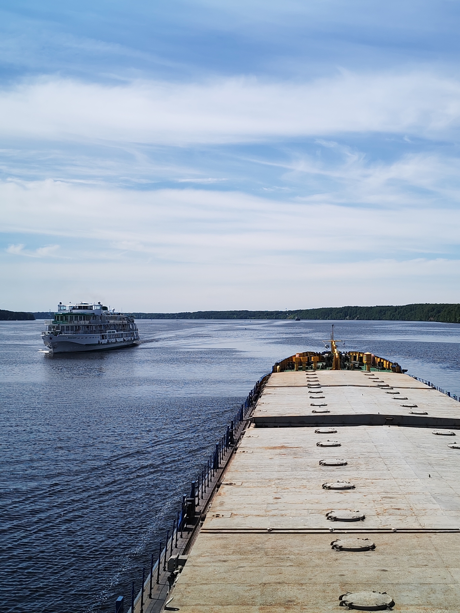 This image captures a wide river scene near the city of Nosovo, Russia, under a blue sky with scattered cirrus and stratocumulus clouds. The foreground is dominated by the flat, weathered deck of a large barge, viewed from the bow looking forward. The barge deck is light grey/beige, featuring multiple rows of circular, bolted hatches, and a blue metal railing visible along its port side. Towards the visible front of the barge, there is a low structure with yellow components, possibly equipment or a small cabin base.
To the left of the barge, in the mid-ground, a multi-decked white passenger ship, likely a river cruise vessel, is observed moving from left to right. The ship exhibits multiple rows of windows and balconies, some with green trim, and a prominent red star emblem on its bow. A distinct wake trails behind the moving passenger ship. The water, a deep blue, shows ripples and patterns from the vessels' movement.
In the far background, dense green forested banks line both sides of the expansive waterway, extending towards the horizon. A very small, dark vessel is faintly visible near the distant right bank. No people are discernible on either the barge or the passenger ship from this perspective. The scene depicts active navigation on a broad river or canal under daylight conditions.